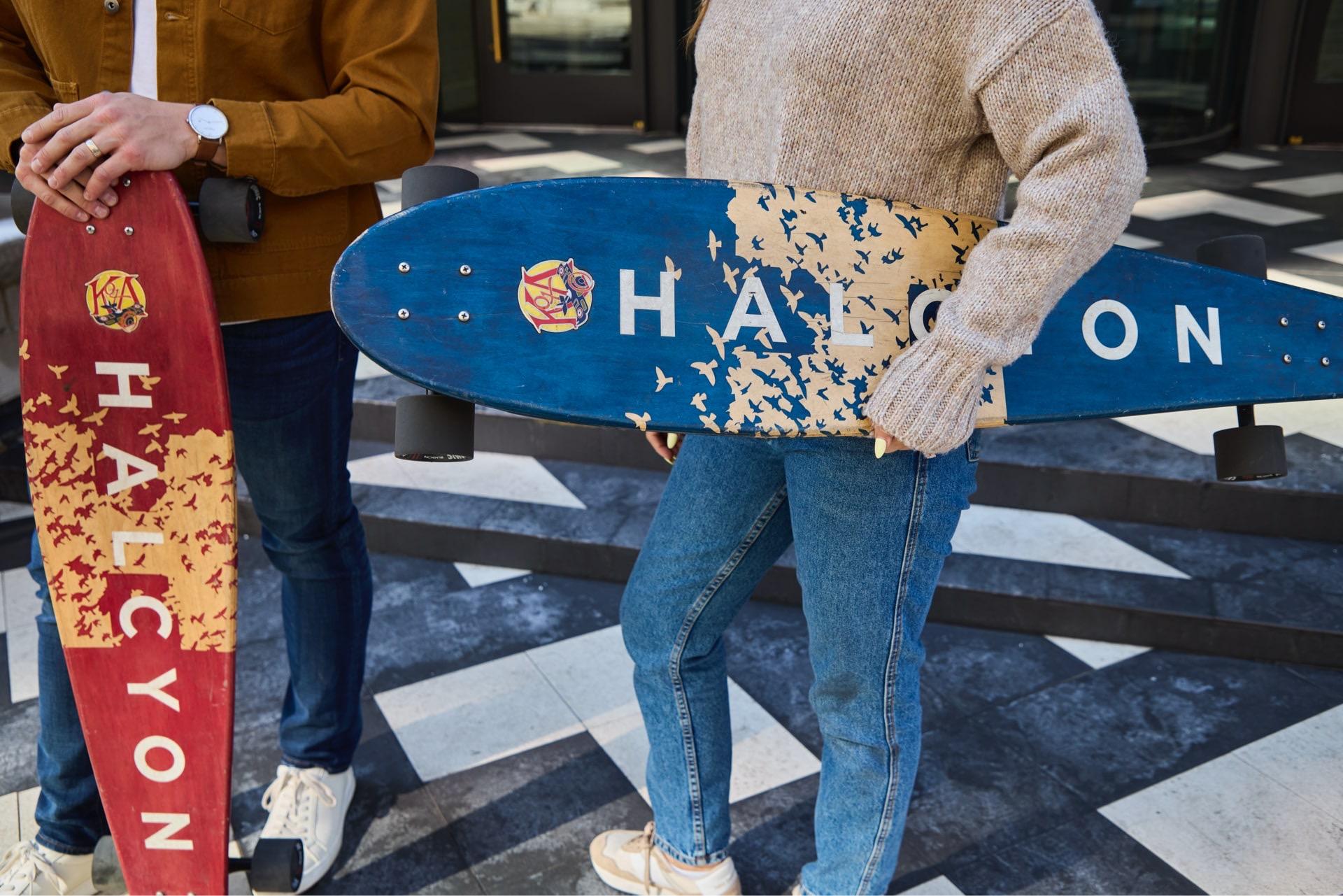 Two people, cropped above the face, hold longboards printed with the Halcyon name in front of a glass entrance, on a black and white tiled plaza.