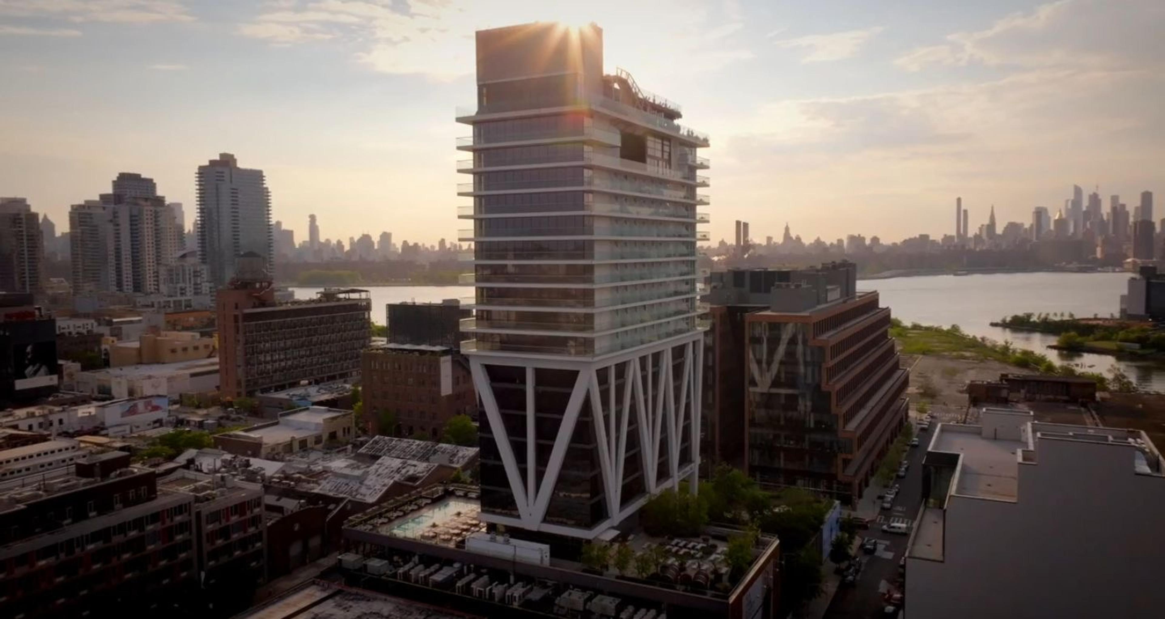 An aerial view of the William Vale's cantilevered tower rising above the Brooklyn skyline, backlit by the setting sun with Manhattan visible across the river.