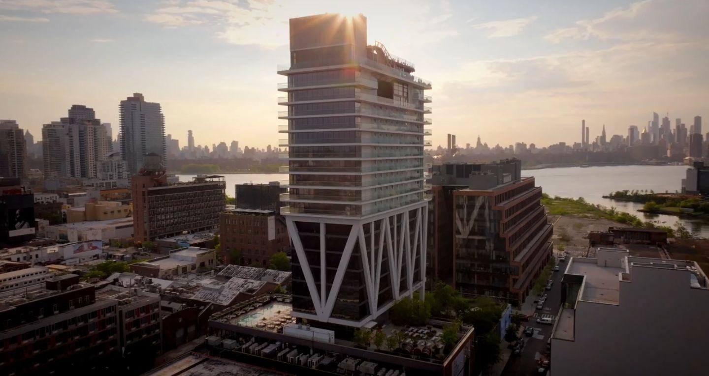 An aerial view of the William Vale's cantilevered tower rising above the Brooklyn skyline, backlit by the setting sun with Manhattan visible across the river.