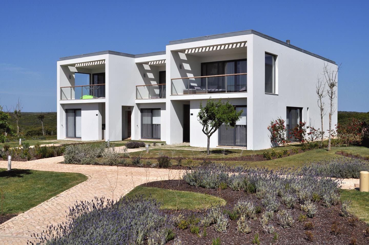 Two-storey white houses with flat roofs beside a paved path, with lavender beds in the foreground and pine trees behind.