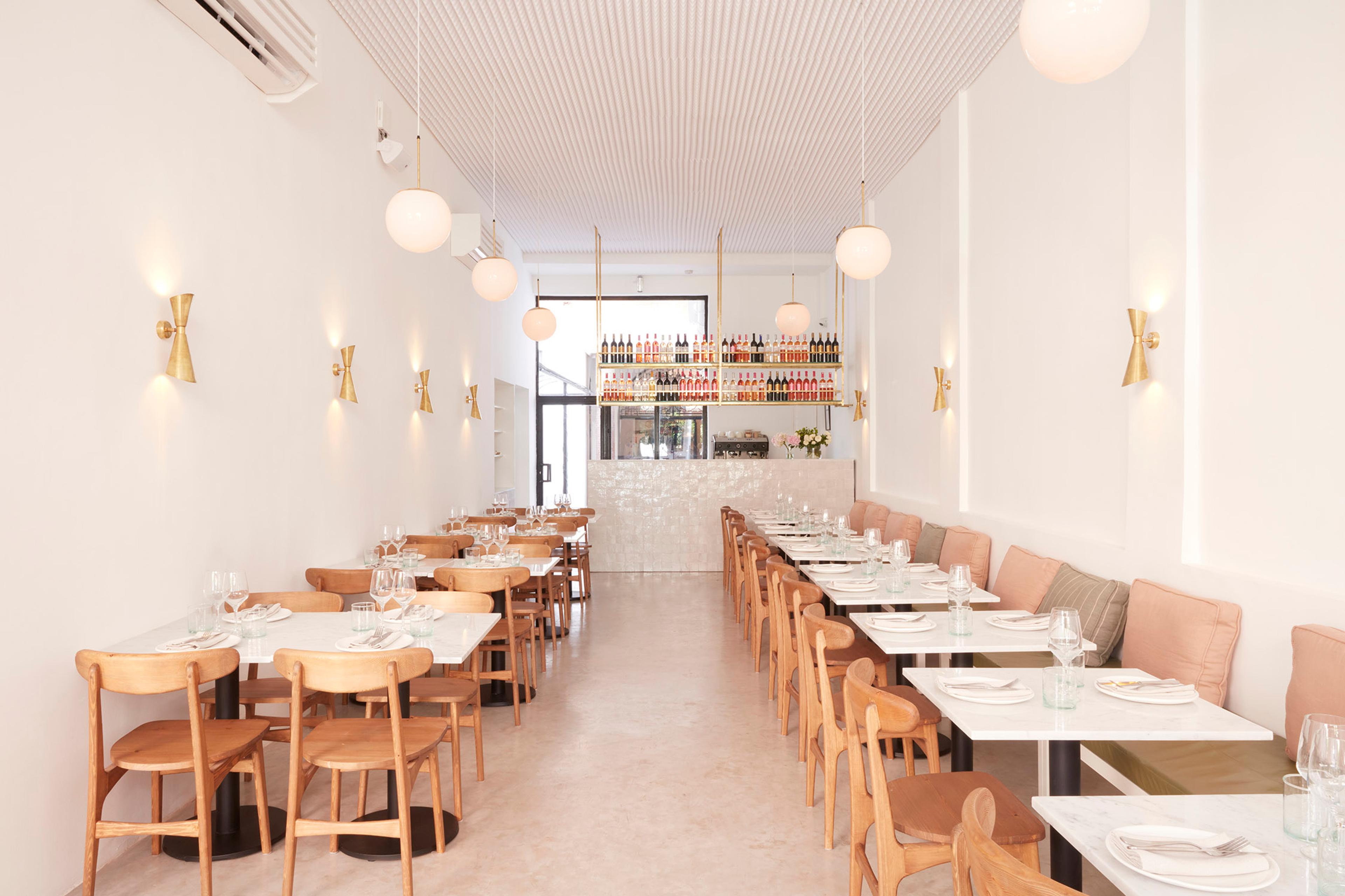 A long, pale dining room with rows of set tables under round wall lamps