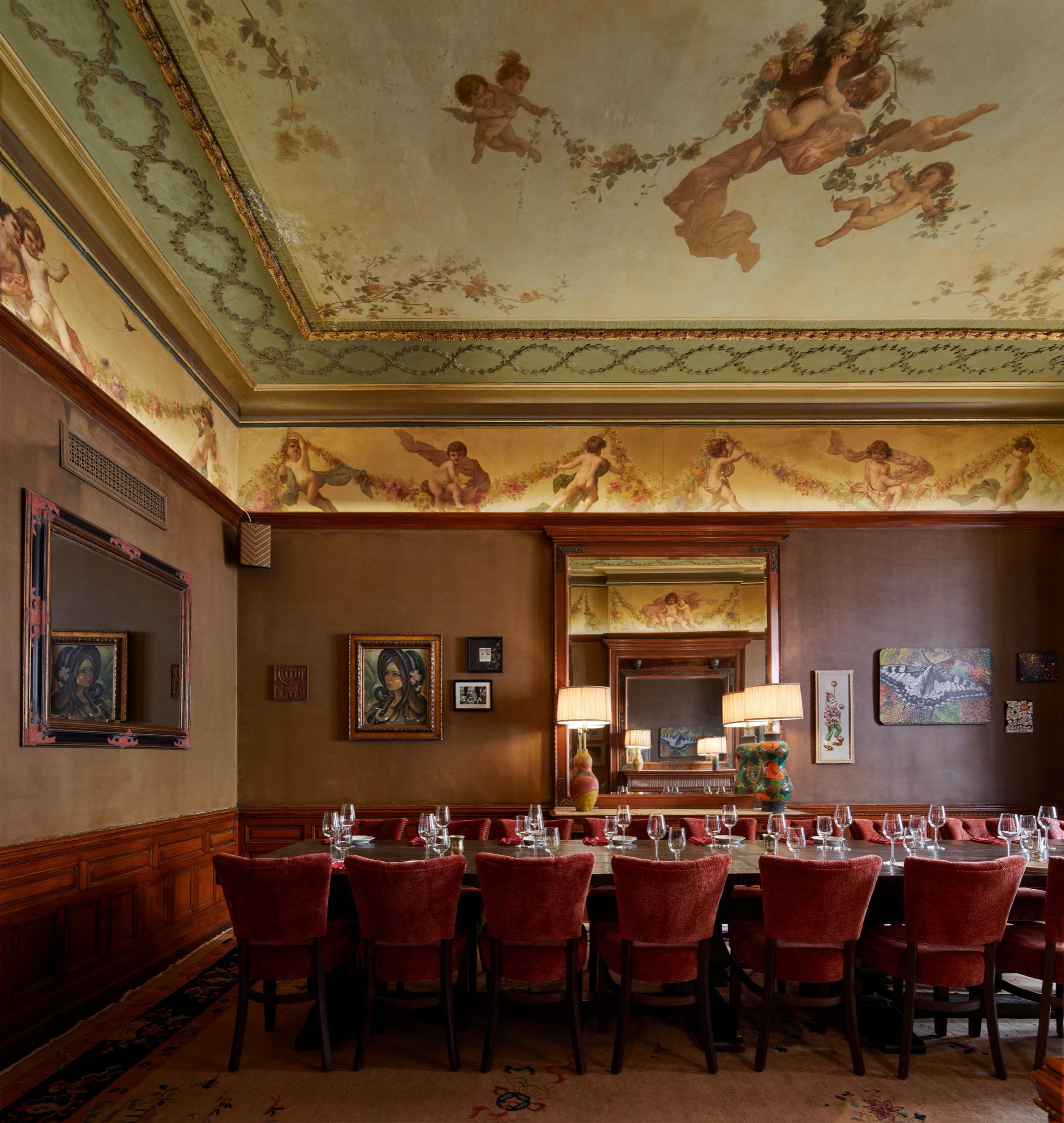 A dining room with a painted cherub ceiling, gilt framed mirrors and artwork, and a long table set with wine glasses surrounded by red chairs.