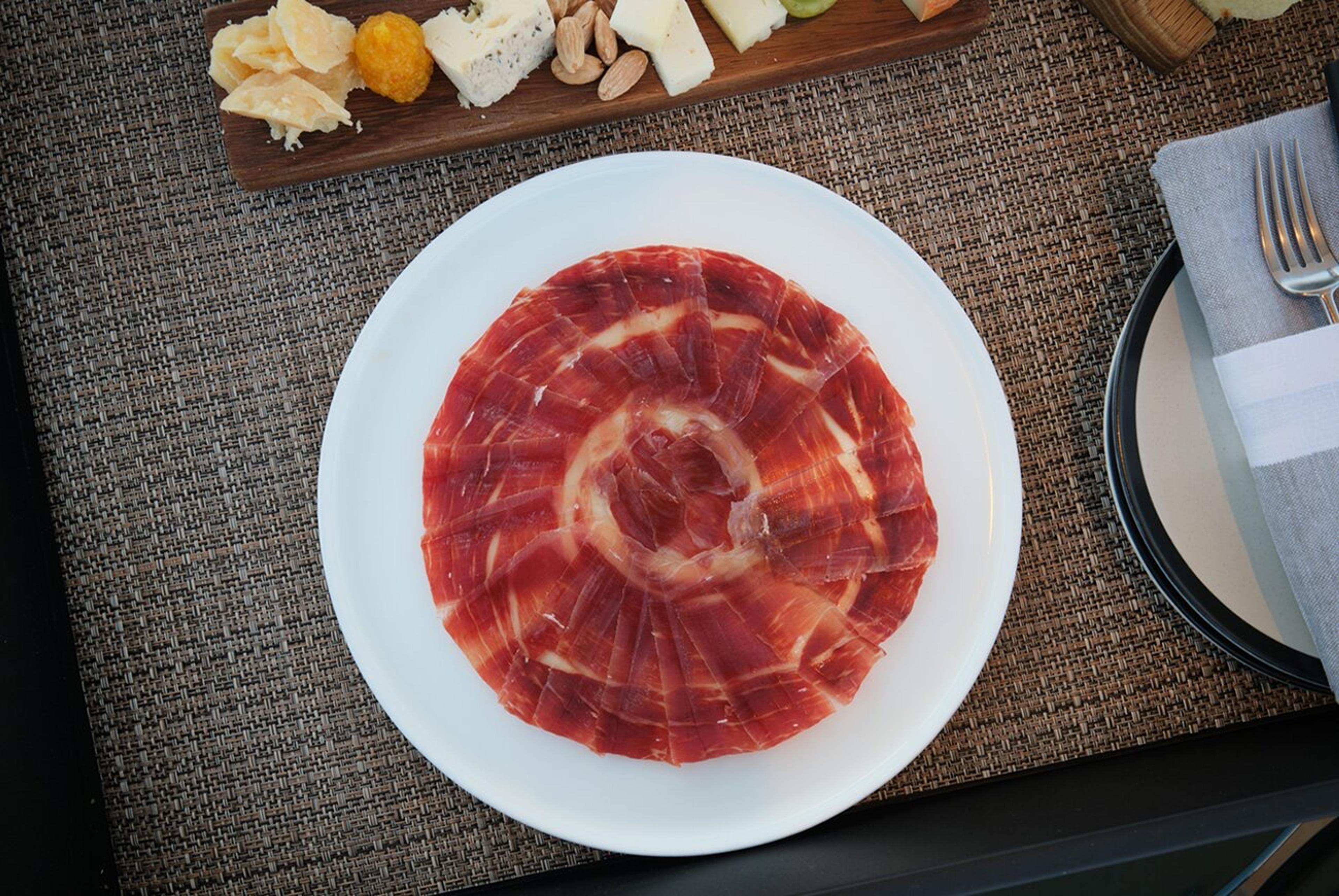 A white plate of thinly sliced cured ham fanned in a circle, beside a wooden board of cheese, dried fruit and almonds, and a folded napkin with a fork.