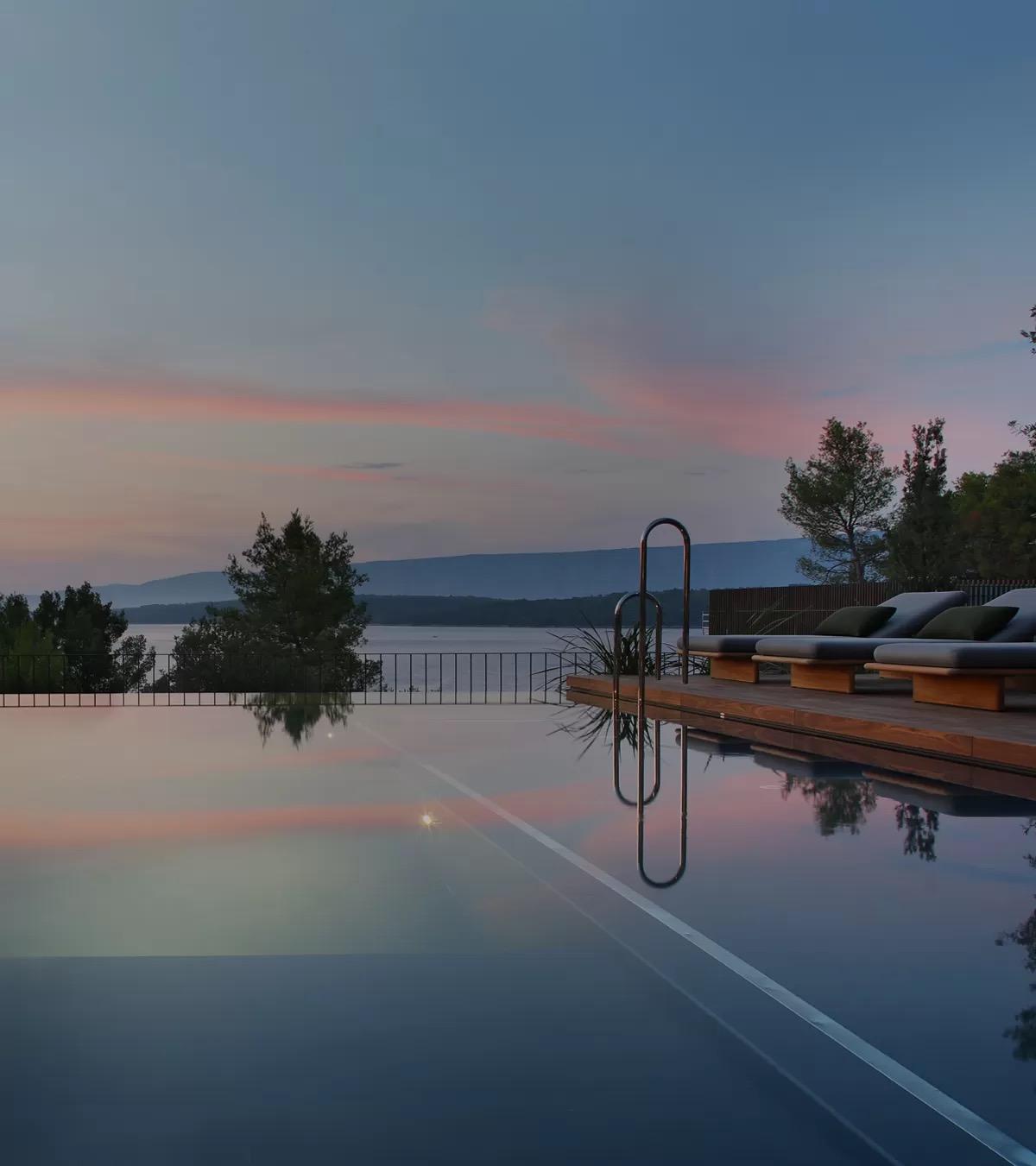 A long infinity pool at dusk, with sunloungers on a timber deck at the right and pine trees and hills beyond the water.