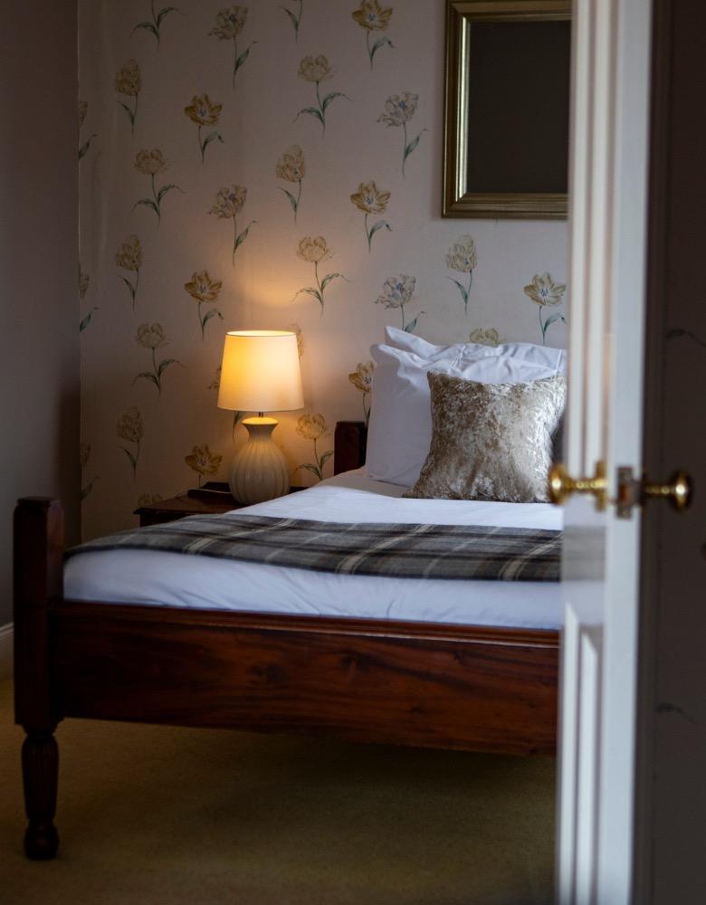 A bedroom with a lamp lit on the bedside table, a patterned headboard cushion and a dark throw across the bed.