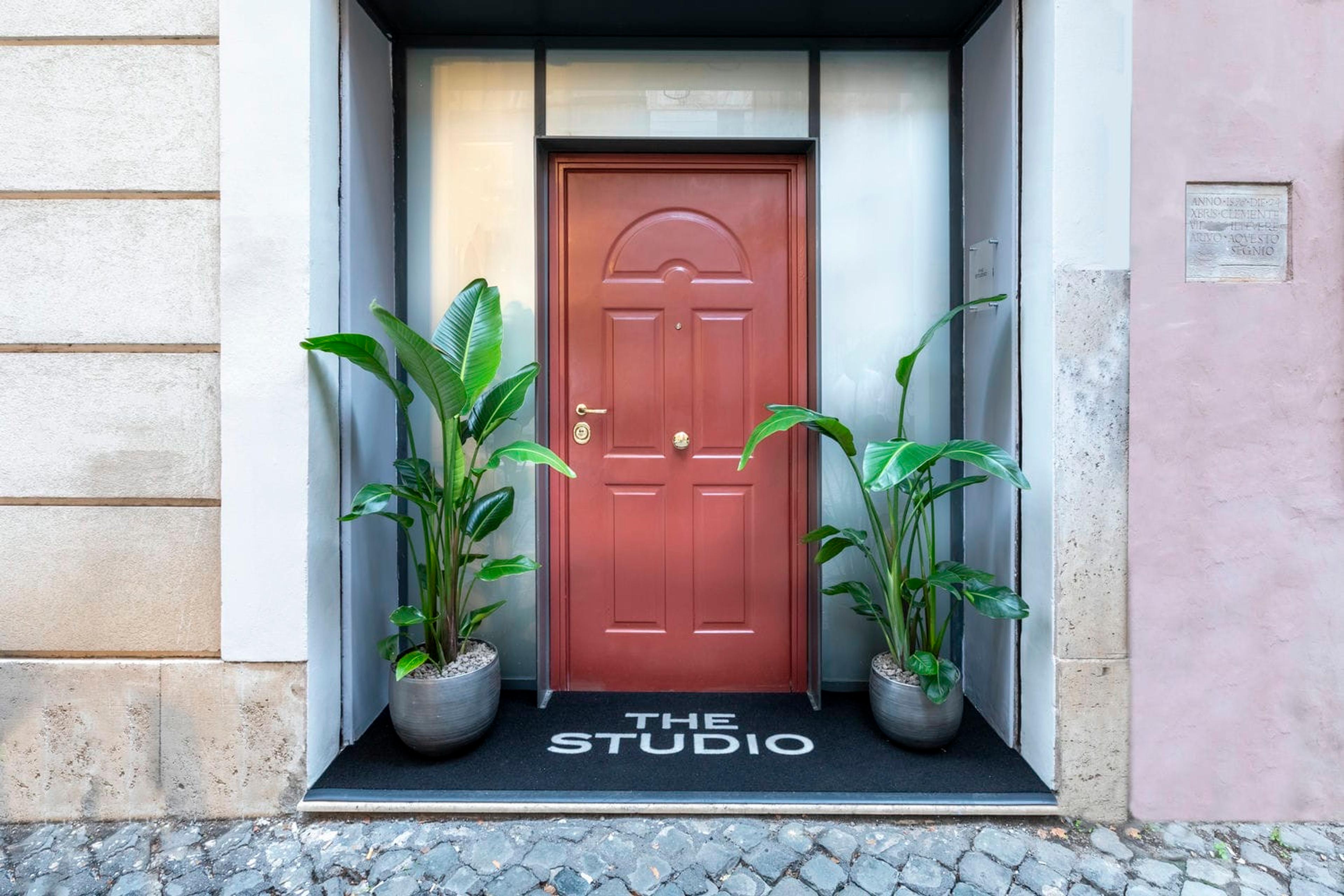 A dark red wooden door in a pale stone doorway, with a potted plant on each side and a black doormat printed with THE STUDIO, on cobbles.