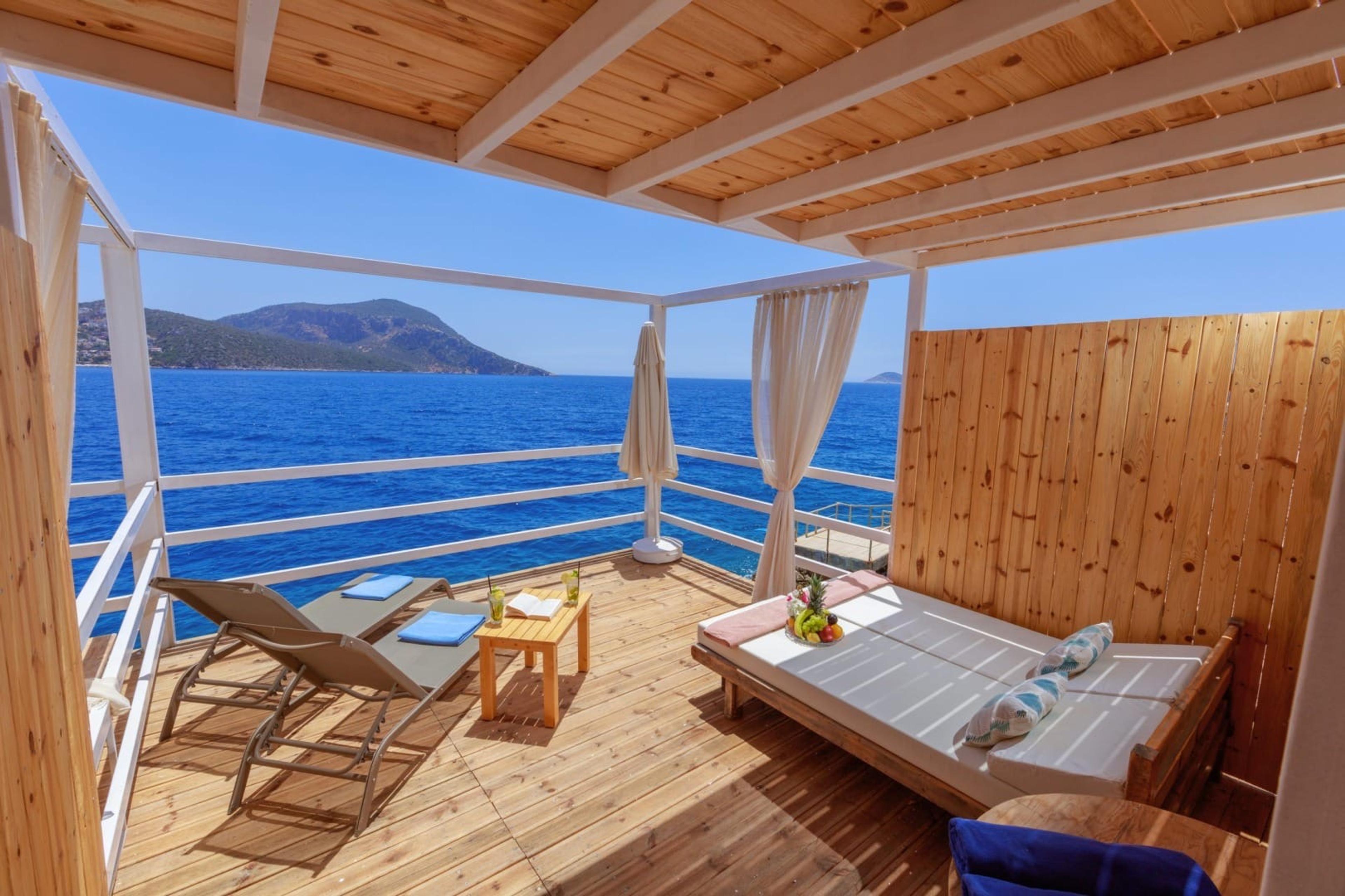 A covered wooden deck with a cushioned daybed and two sun loungers, looking out over open sea to distant hills.