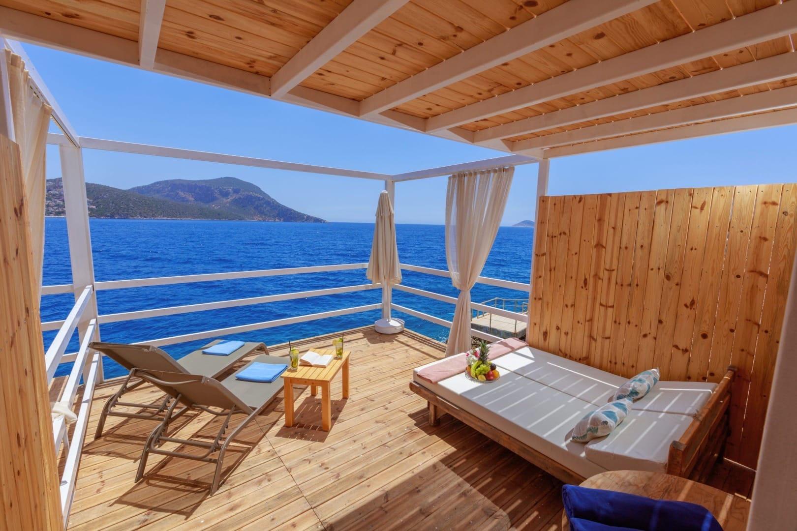 A covered wooden deck with a cushioned daybed and two sun loungers, looking out over open sea to distant hills.