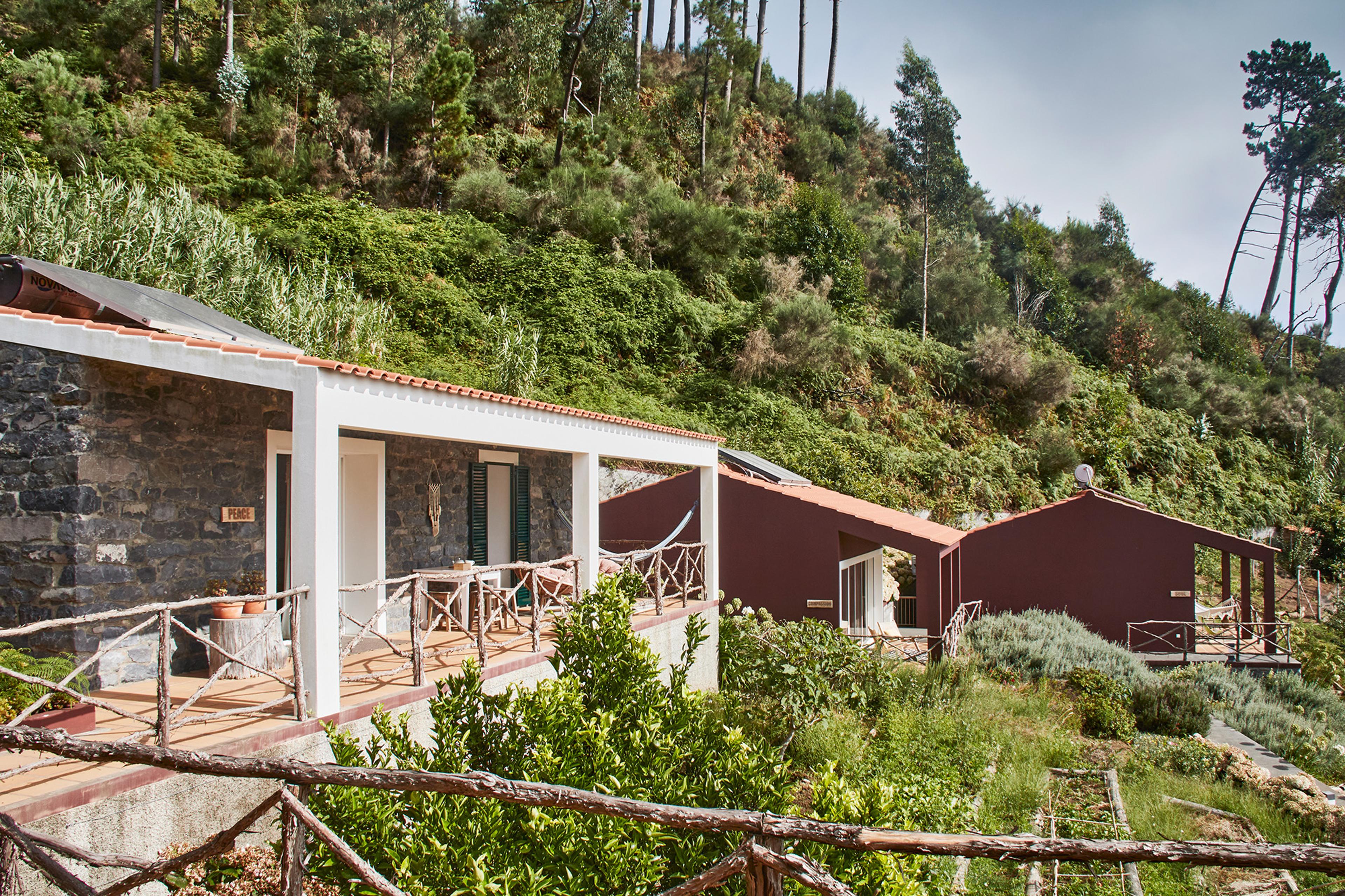 A stone building with a white-columned covered terrace and a hammock, beside dark red outbuildings set into a wooded hillside.