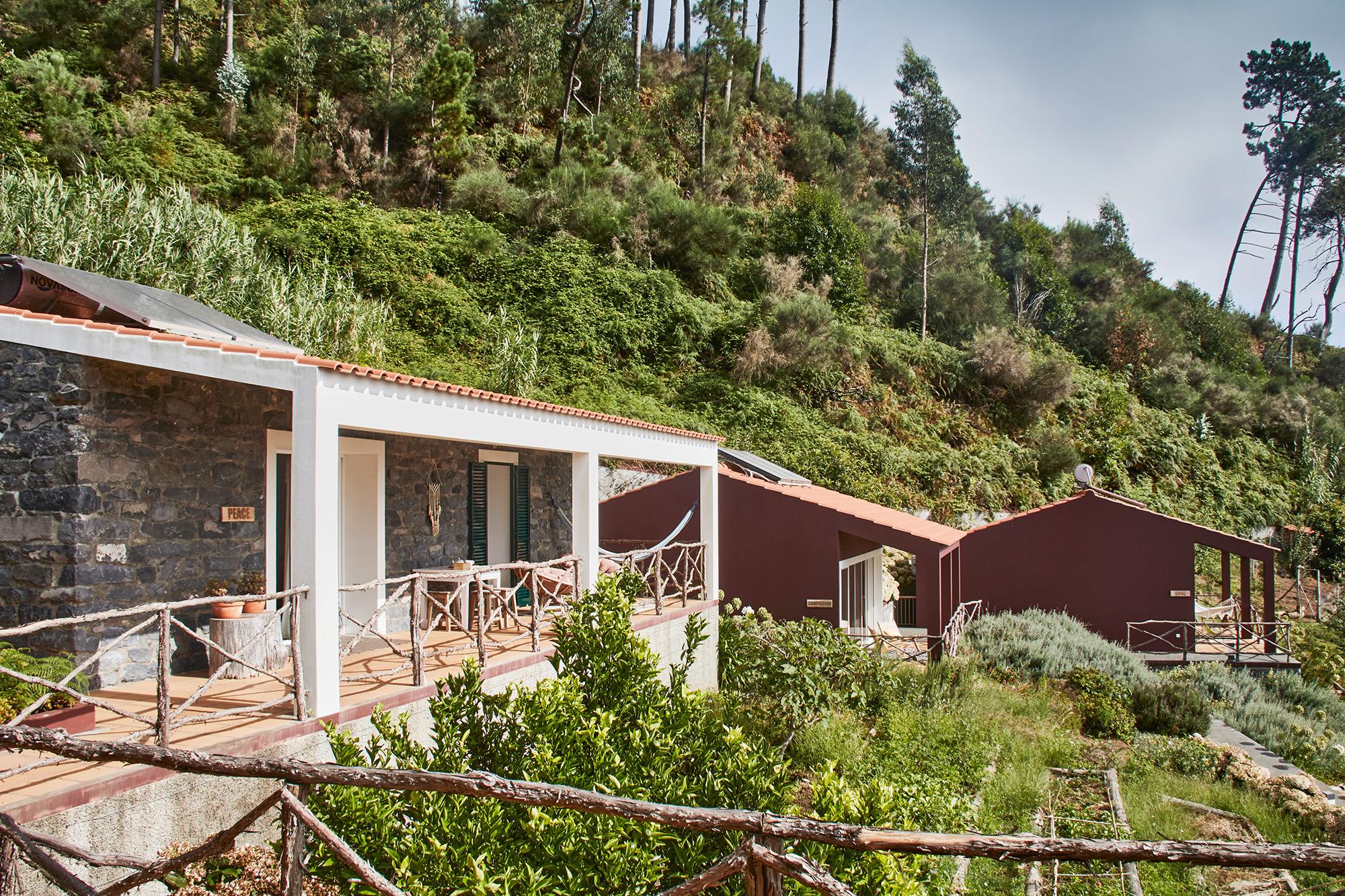 A stone building with a white-columned covered terrace and a hammock, beside dark red outbuildings set into a wooded hillside.