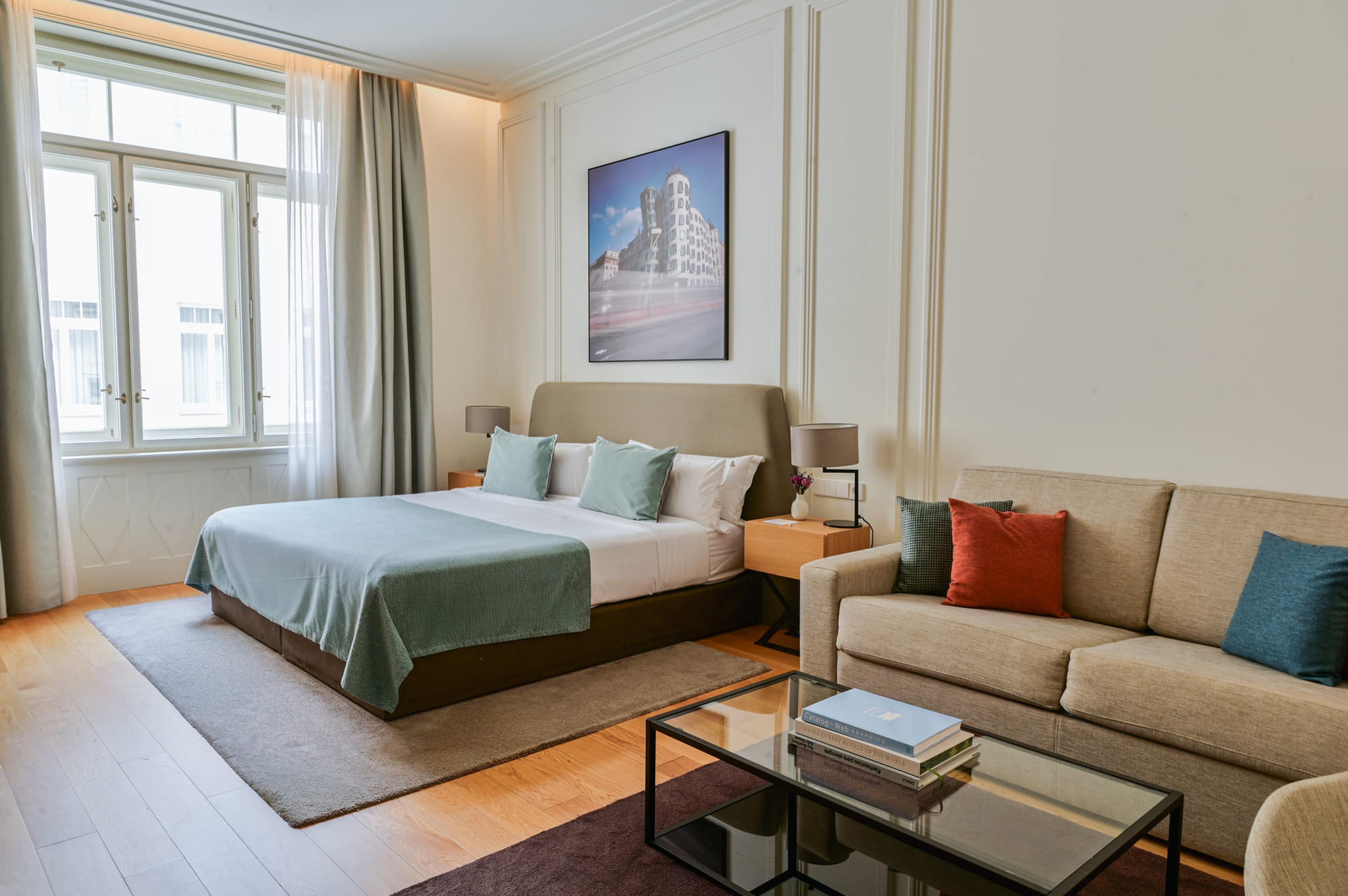 A bedroom with a grey-green bed, a beige sofa with orange and red cushions, and a painting of the Dancing House in Prague above the desk.