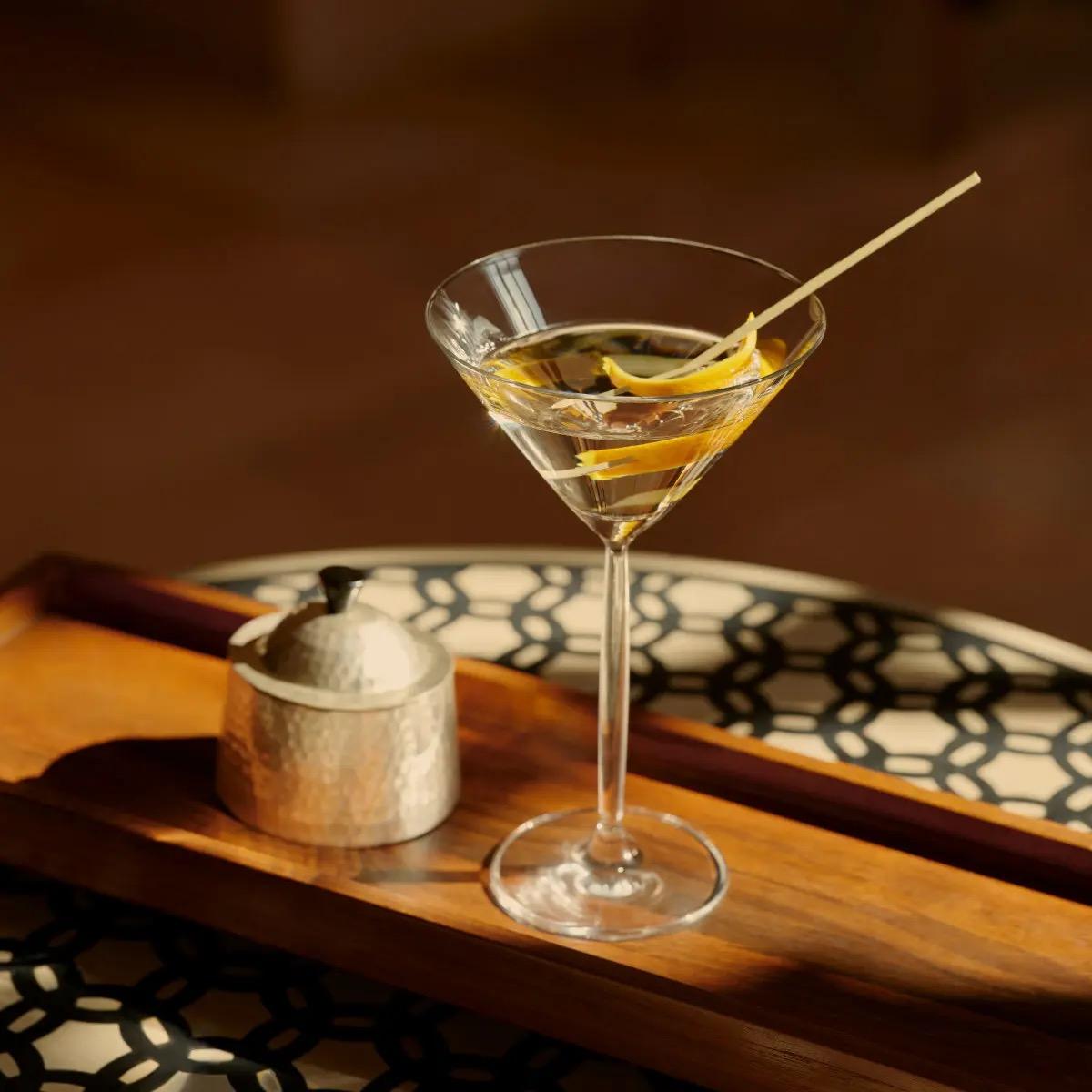 A martini glass with a long citrus twist and a swizzle stick, on a wooden tray beside a small lidded jar.