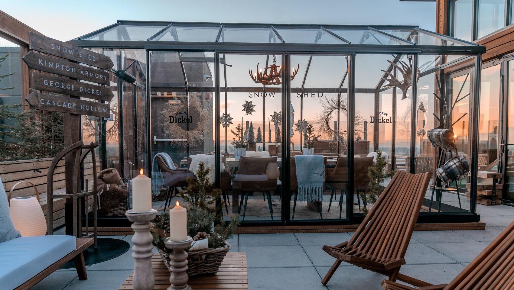 A glass-walled rooftop structure lit up at dusk, with lanterns, candles and wooden chairs on the terrace in front of it and a wooden signpost reading Snow Shed and Kimpton Armory.