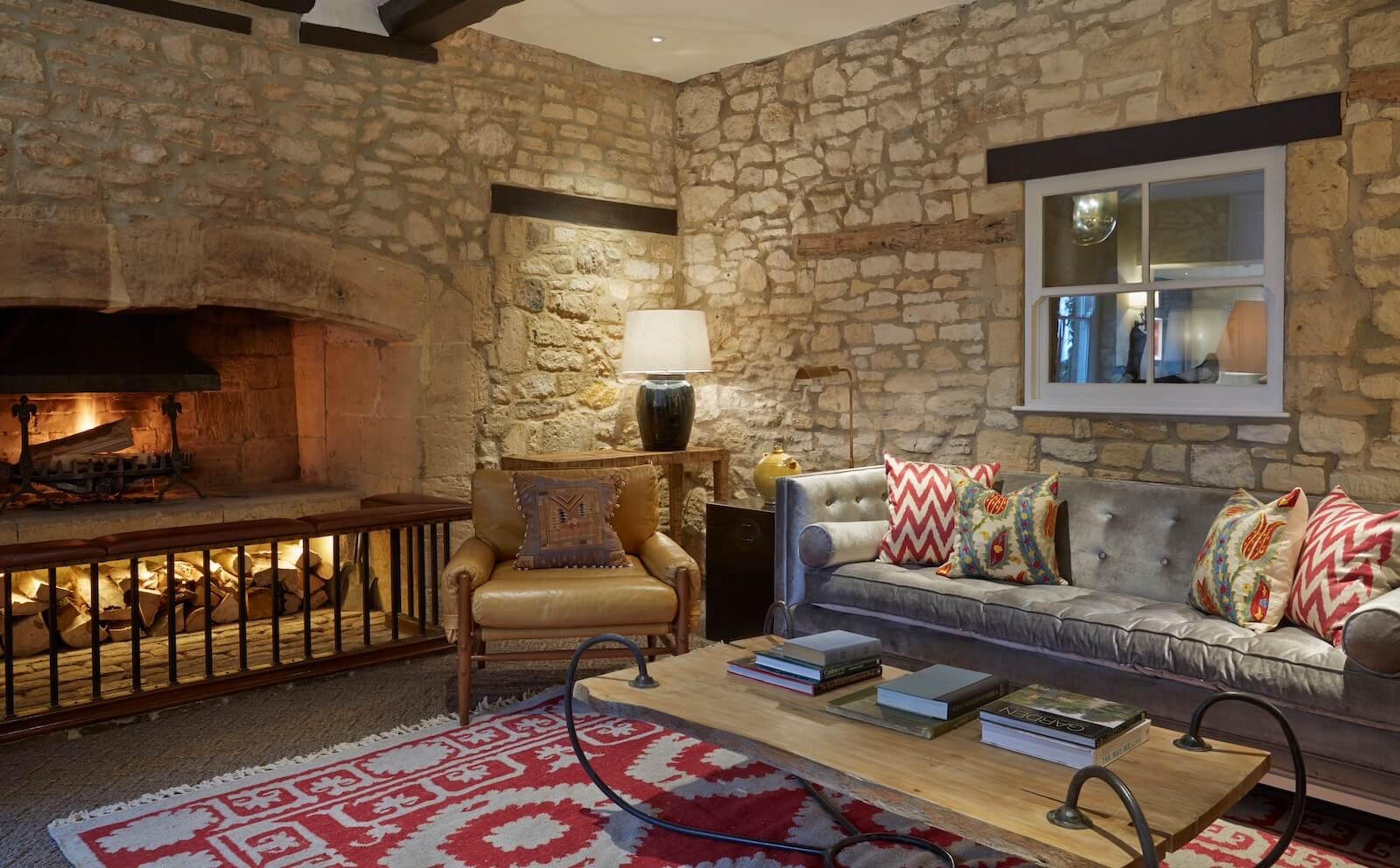 A stone-walled lounge with a large open fireplace, a grey tufted sofa with patterned cushions, and a red and white patterned rug.