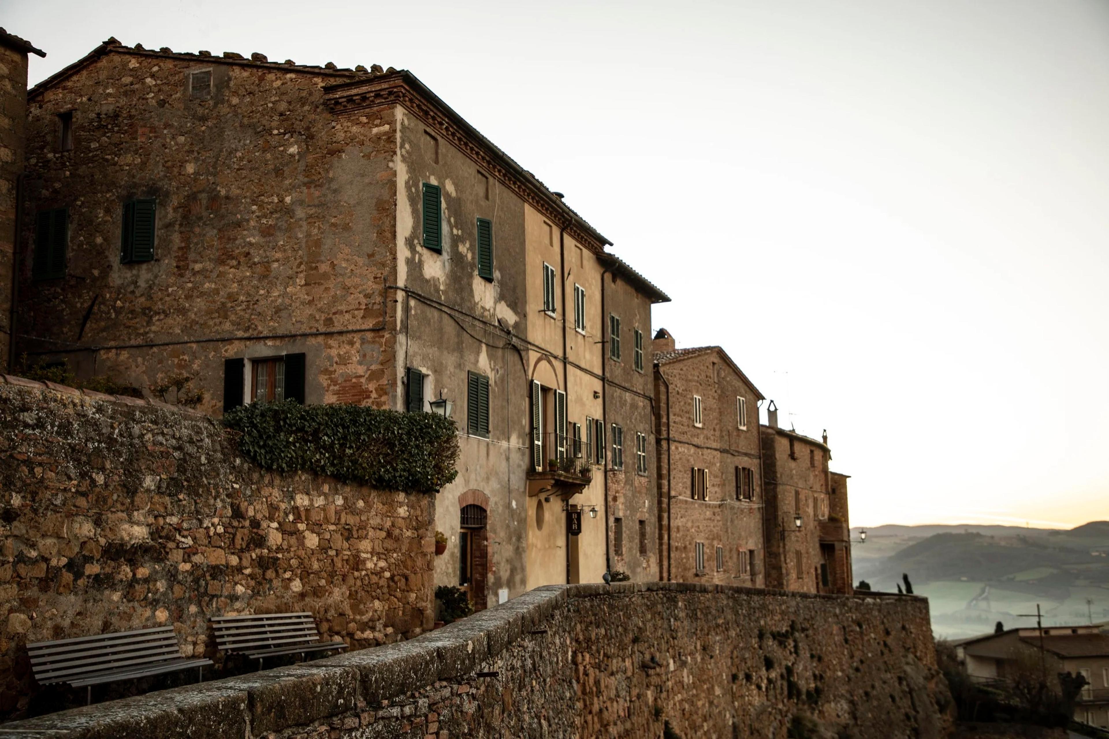 A row of stone townhouses along a walled hillside street at dusk, with two benches and hills visible beyond.