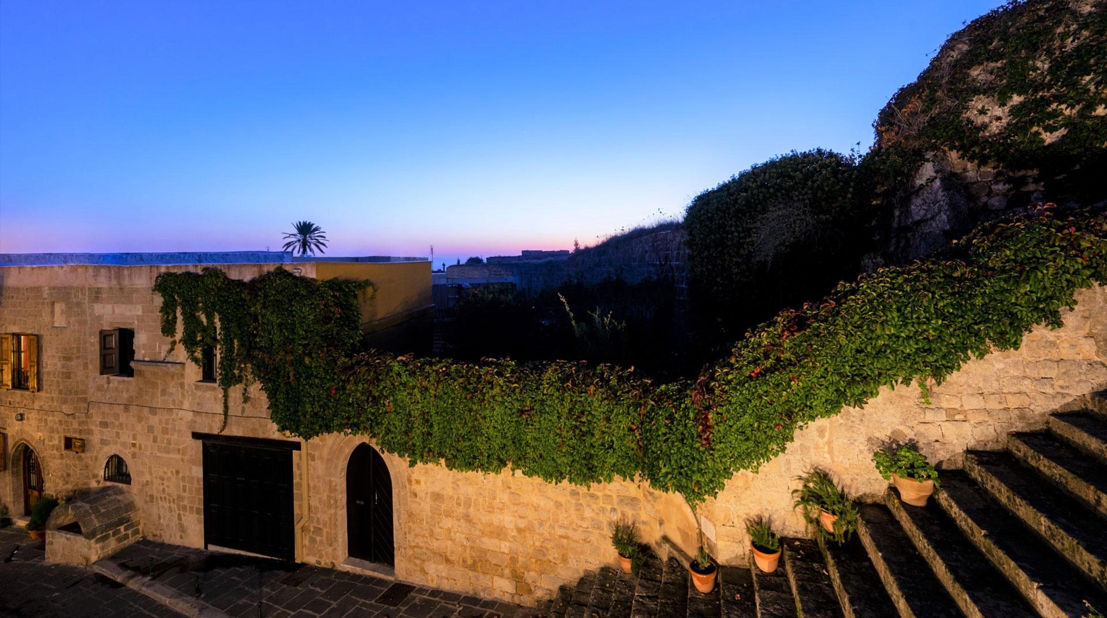 A stone building with an ivy-covered boundary wall and stone steps, photographed at dusk under a palm tree.