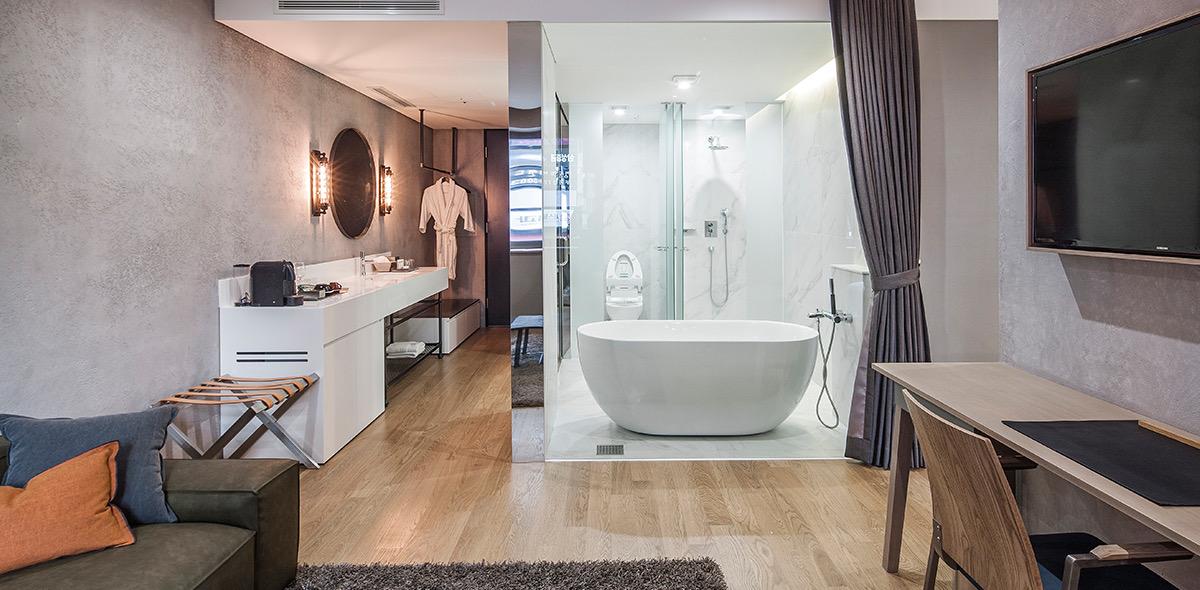A freestanding white bathtub behind a glass partition, with a sink counter, a hanging bathrobe and a round mirror in the room beyond.