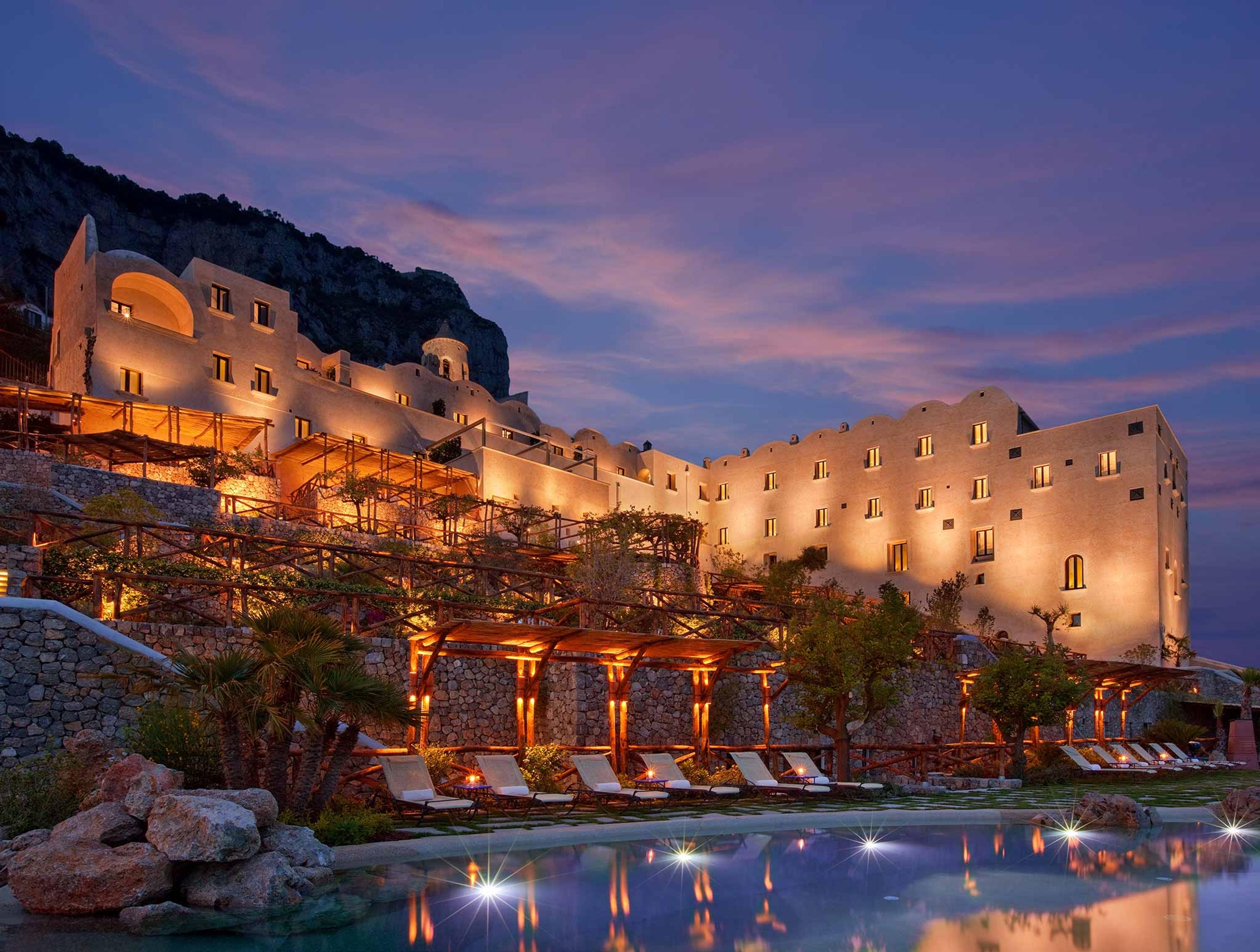 the monastery lit gold against a pink dusk sky, its infinity pool and loungers in the foreground at the base of the cliff