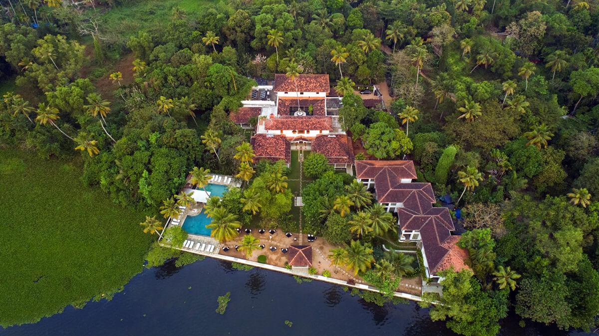 An aerial view of a resort with red tiled roofs set among palm trees beside a lake, with a swimming pool and sun loungers near the water's edge.