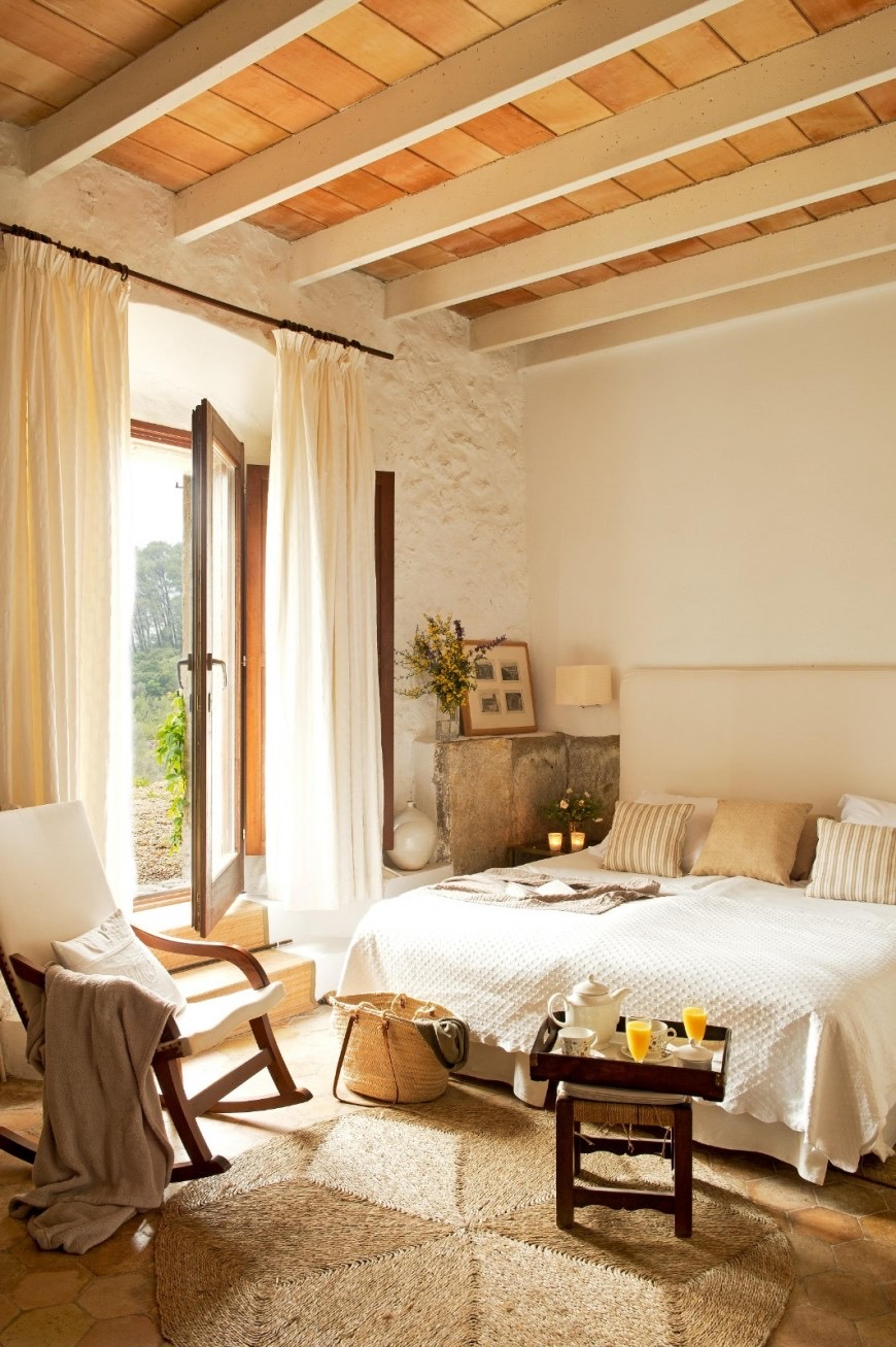A breakfast tray of coffee and orange juice sits on a stool by a bed with striped cushions, the door open to greenery outside and a rocking chair draped with a throw.