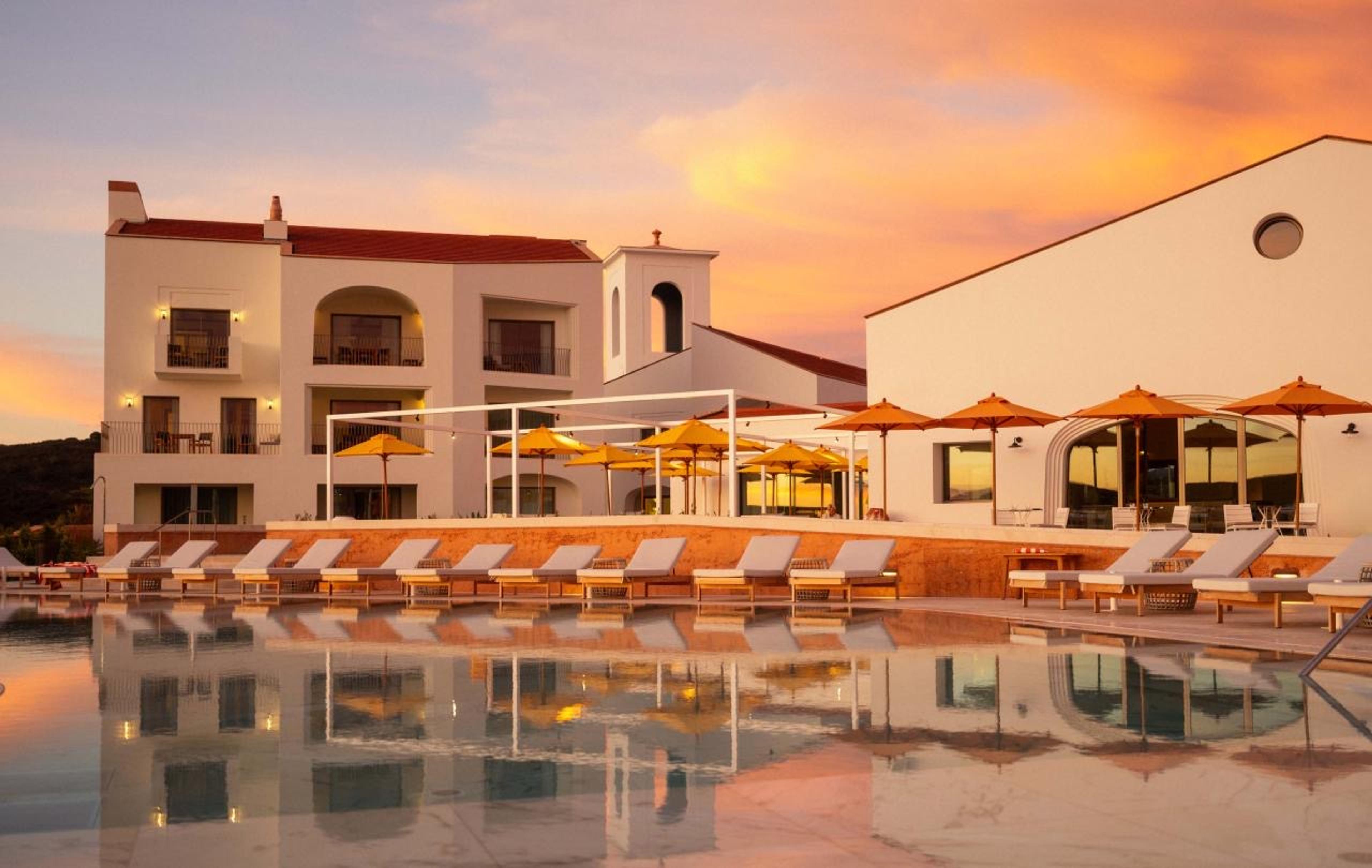 A row of whitewashed buildings and a pool terrace with orange umbrellas at sunset, the pool reflecting a pink and orange sky.