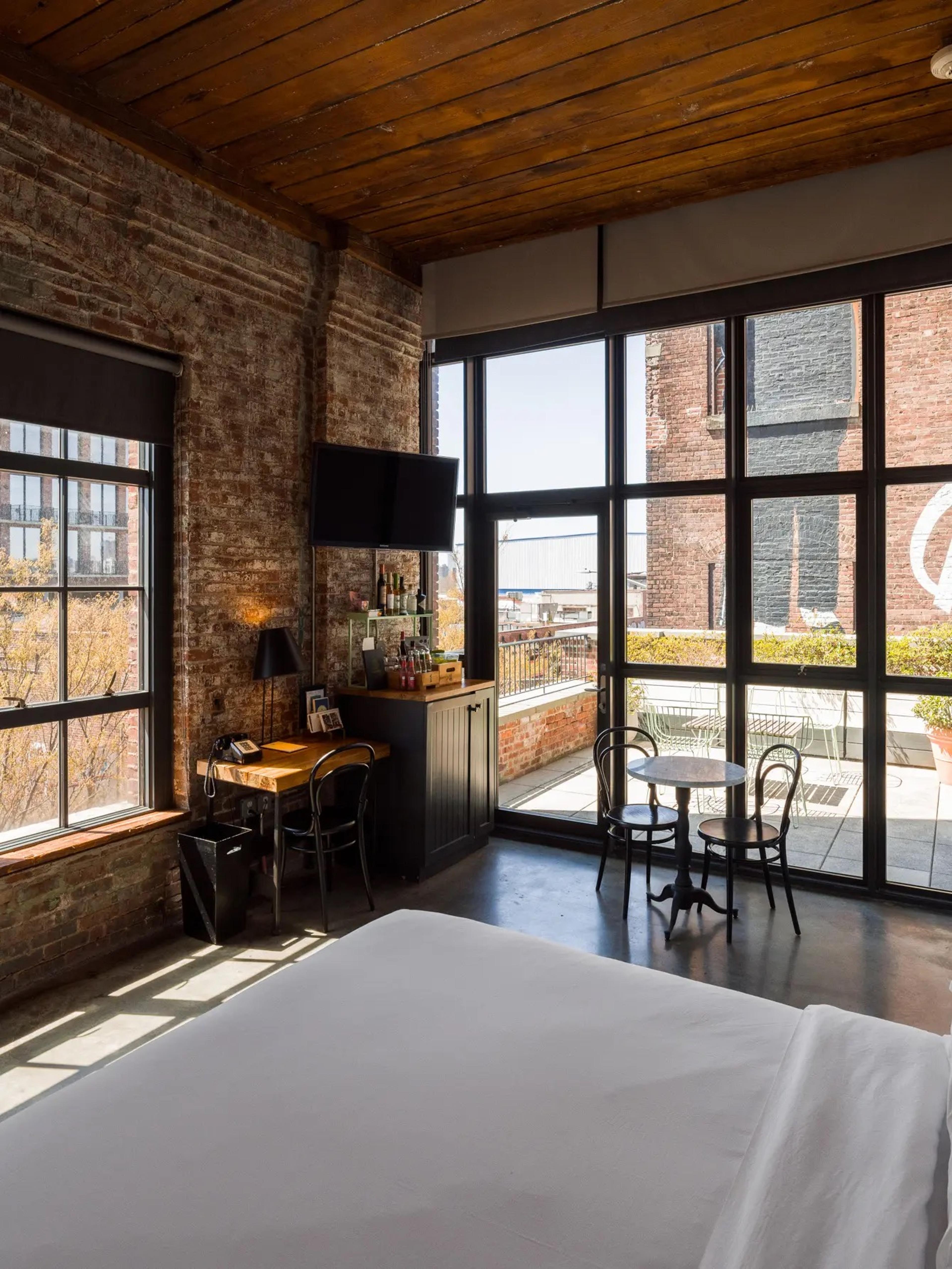 A hotel room with brick walls, a desk and television, opening onto a private rooftop terrace with outdoor furniture and a neighbouring brick building.