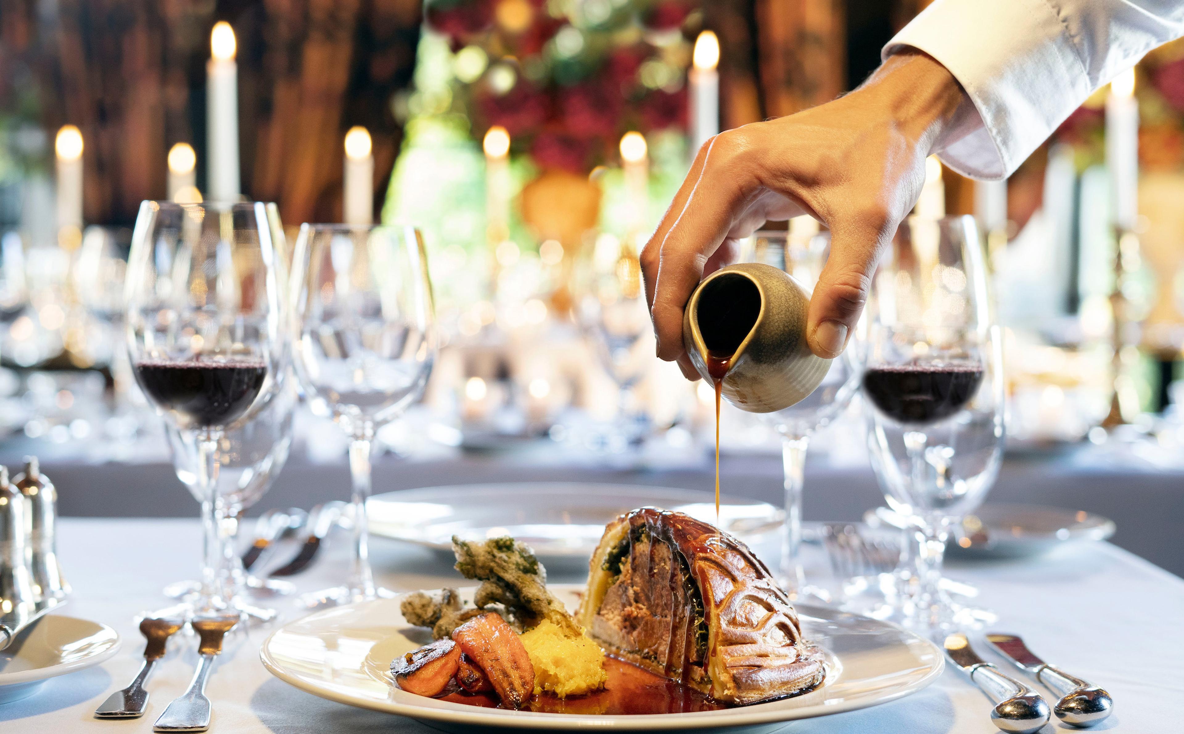 a hand pours sauce over a plated main course of wrapped meat and vegetables on a table set with wine glasses and candles