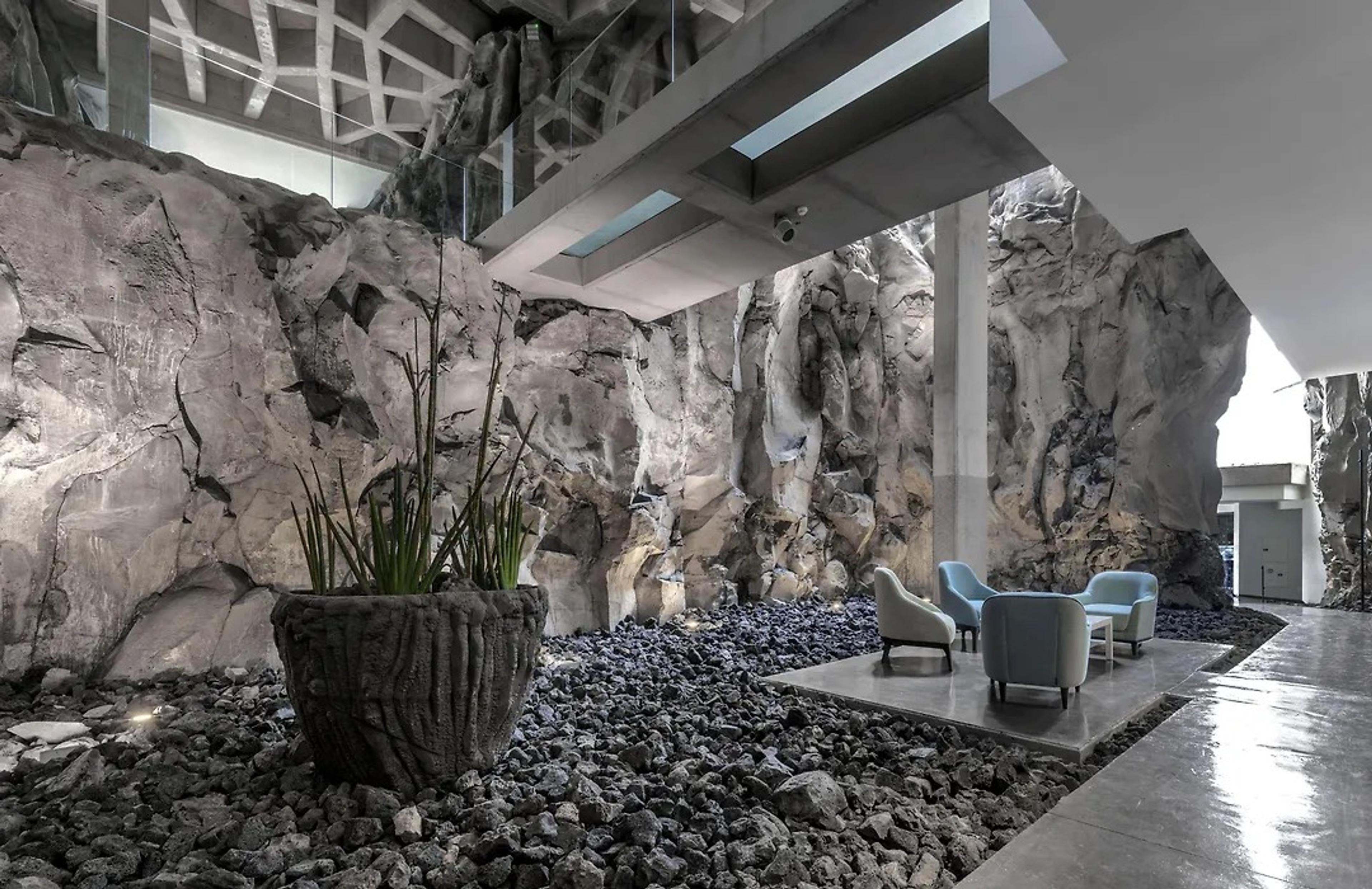 A lobby of rough volcanic rock walls with a gravel floor, a planted urn and blue armchairs