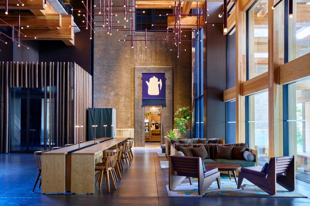 A long hotel lounge with a communal wood table and chairs down one side, low armchairs and a sofa around a coffee table on the other, under a ceiling hung with string lights and a lit sign shaped like a kettle.