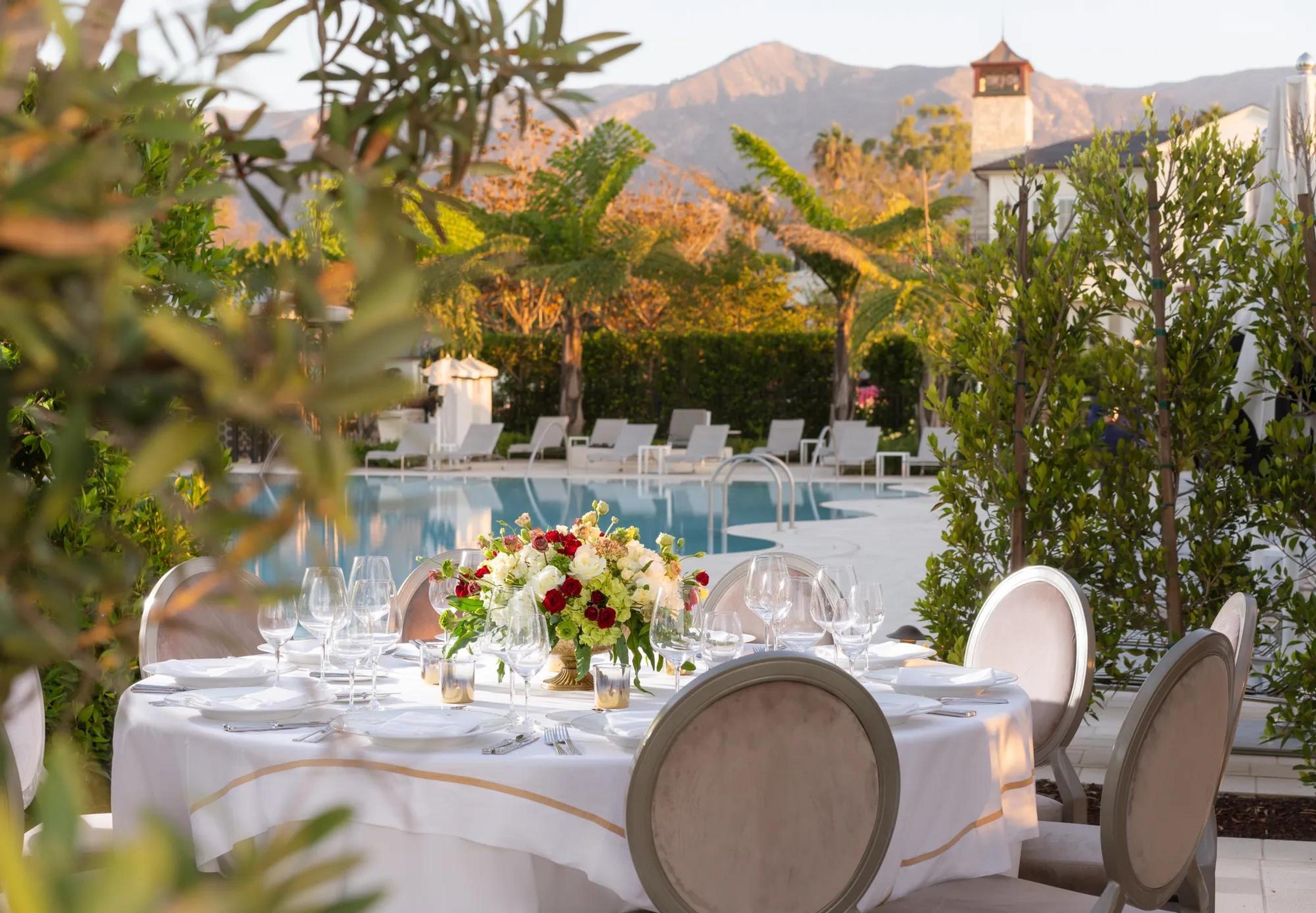 A round table set with flowers beside a swimming pool, with mountains in the background.