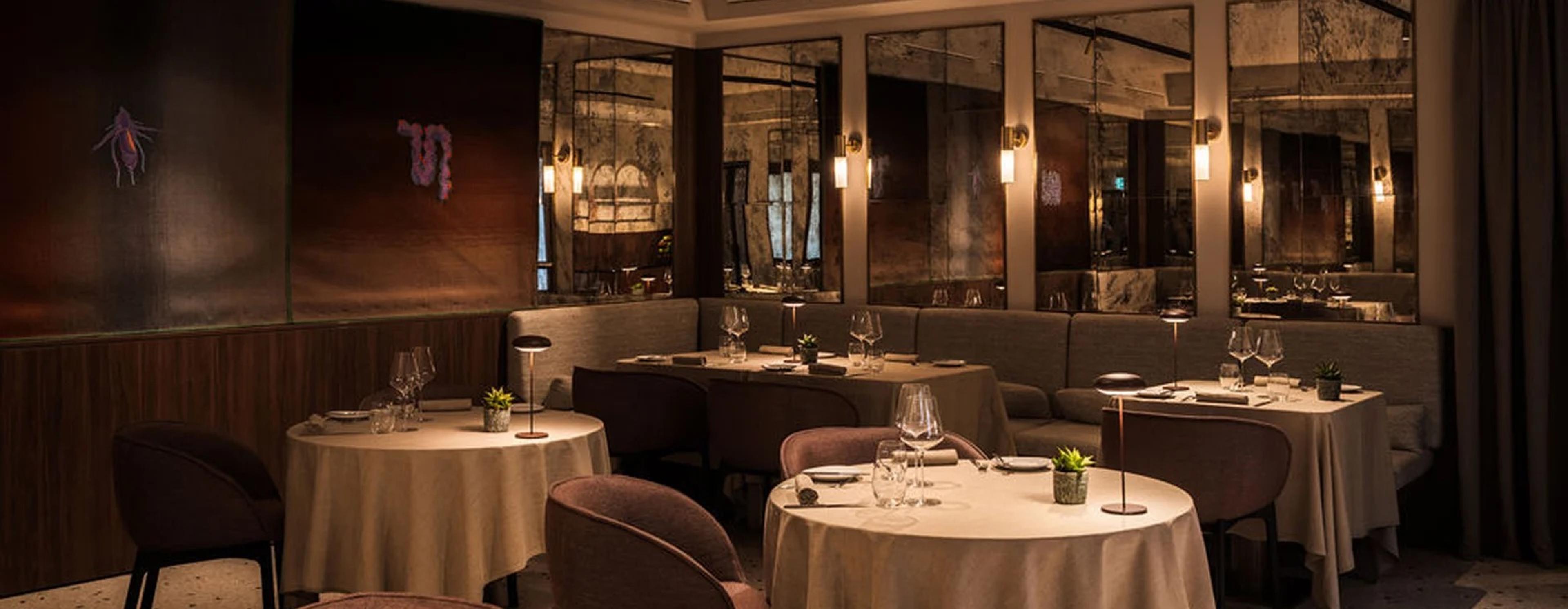 An empty restaurant dining room with round tables set for dinner, banquette seating, and mirrored wall panels.
