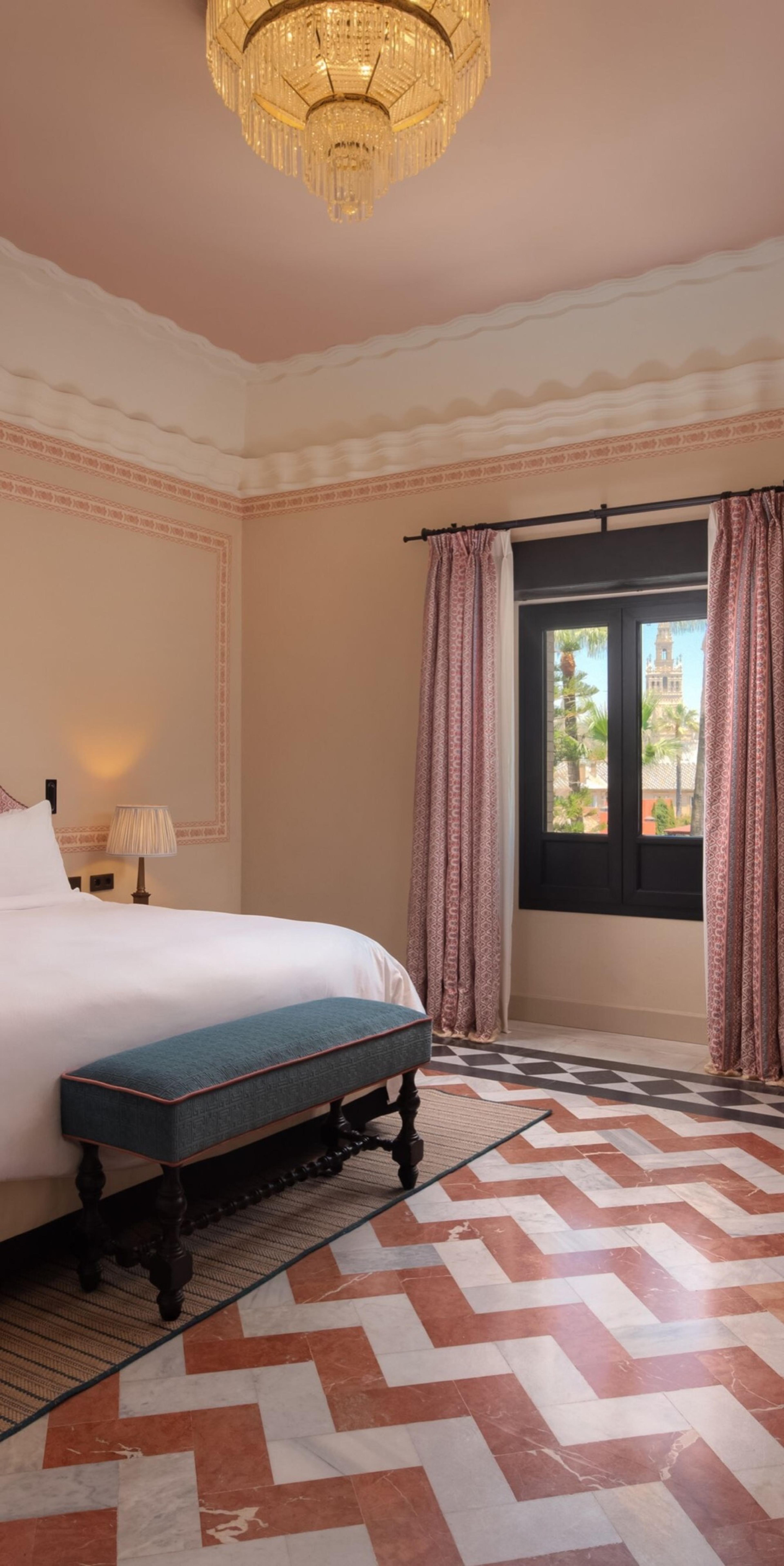 A twin bedroom under a carved wood coffered ceiling, with a crystal chandelier, diamond-patterned headboards and floral curtains open to a balcony door.