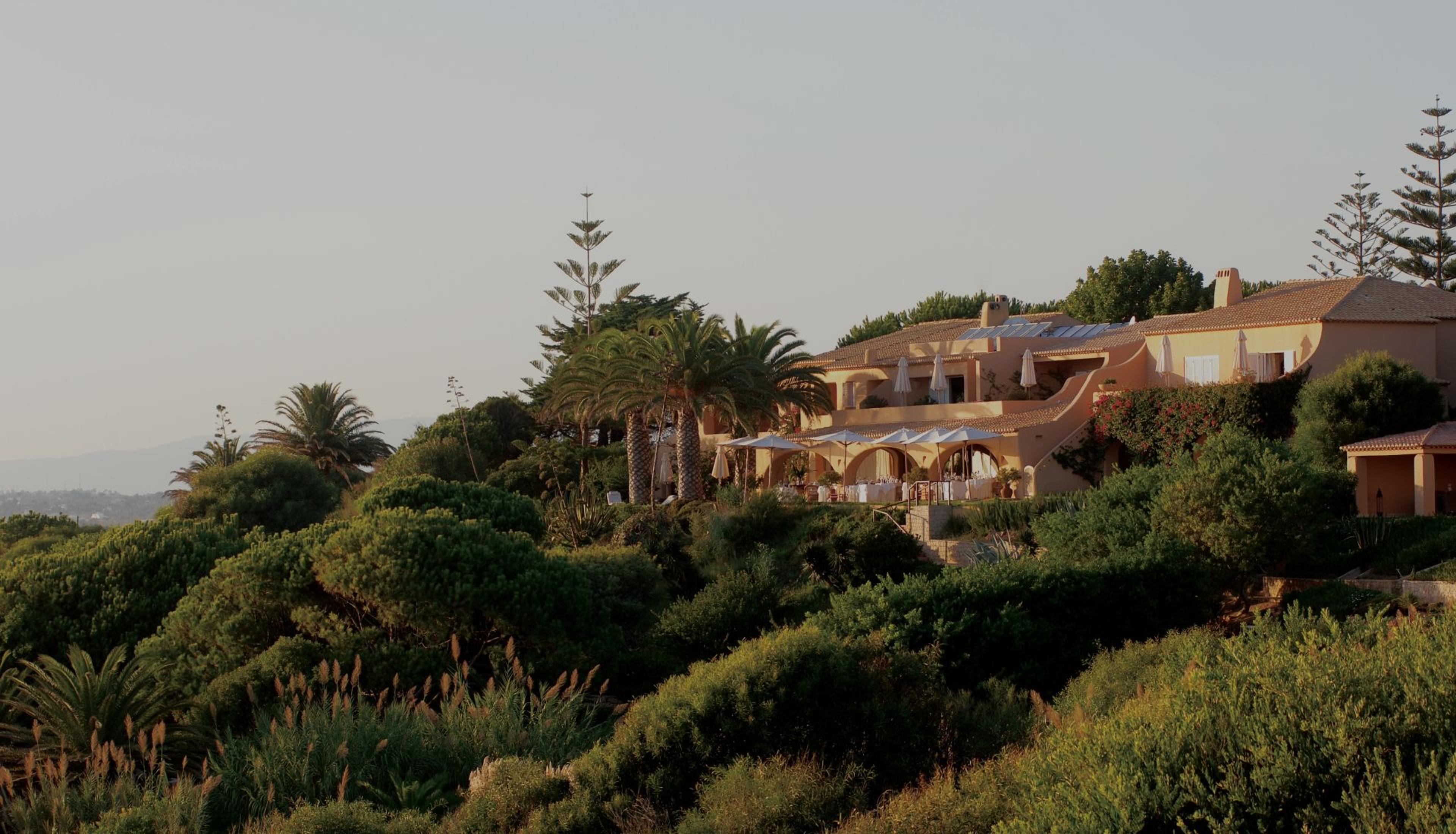 A cluster of ochre buildings with tiled roofs set among pine and palm trees on a hillside, lit by late afternoon sun.