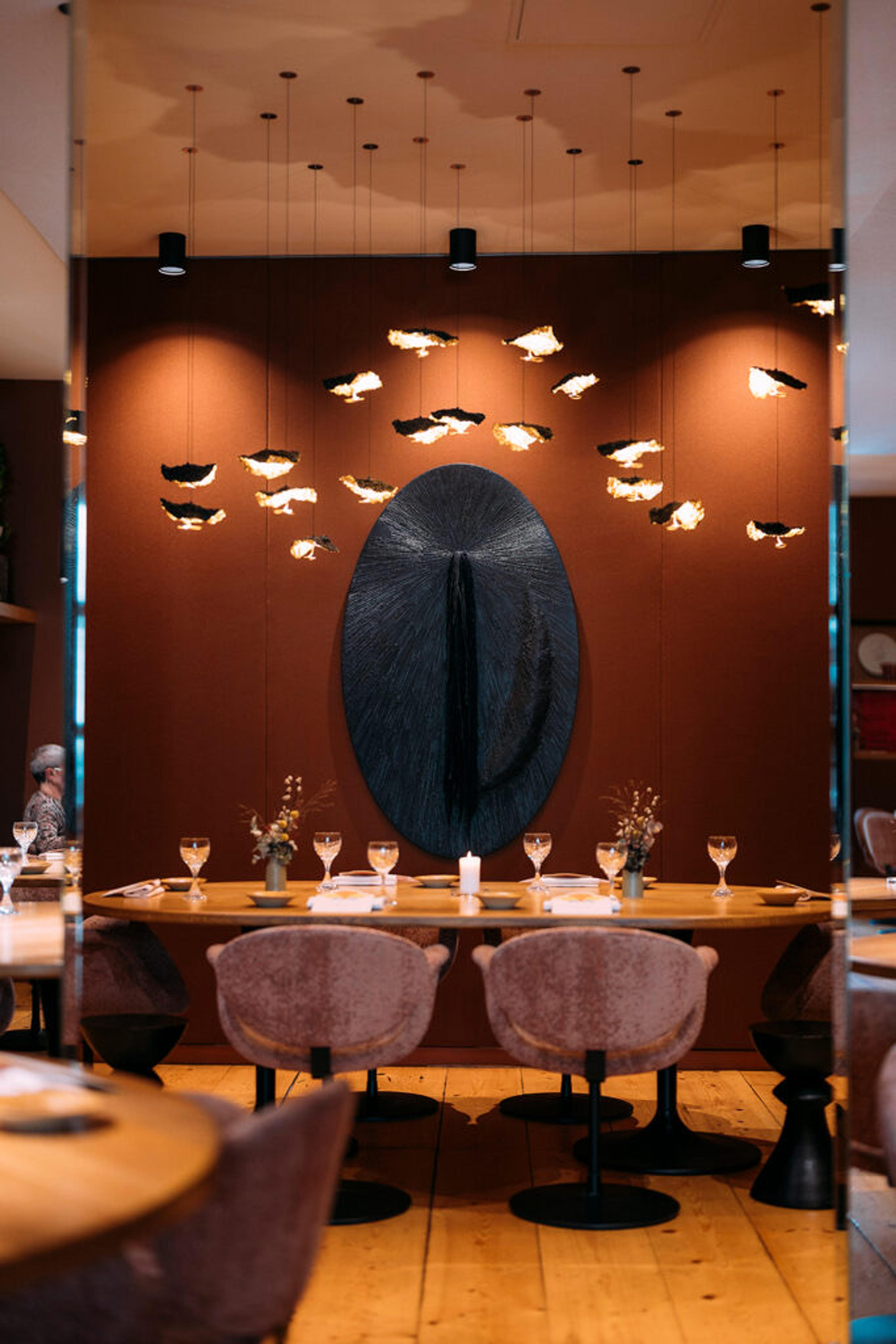 A dining room with terracotta walls, a dark sculpture on the far wall and tables laid with glasses.
