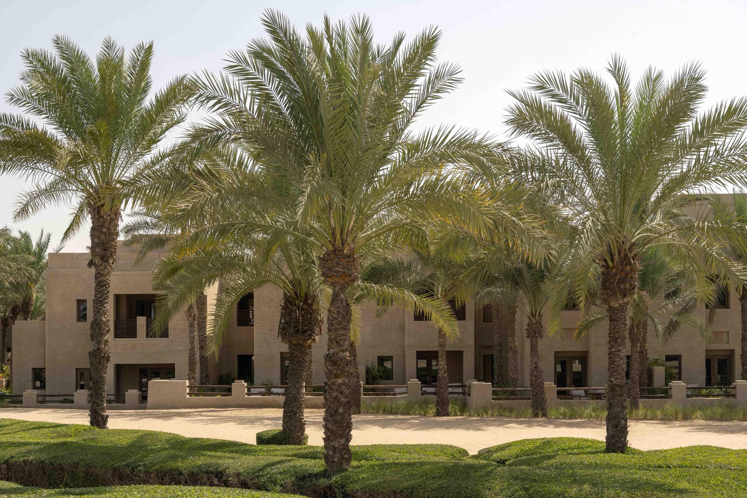 A low sand-colored guest wing with balconies and arched windows, seen through a screen of date palms in daylight.