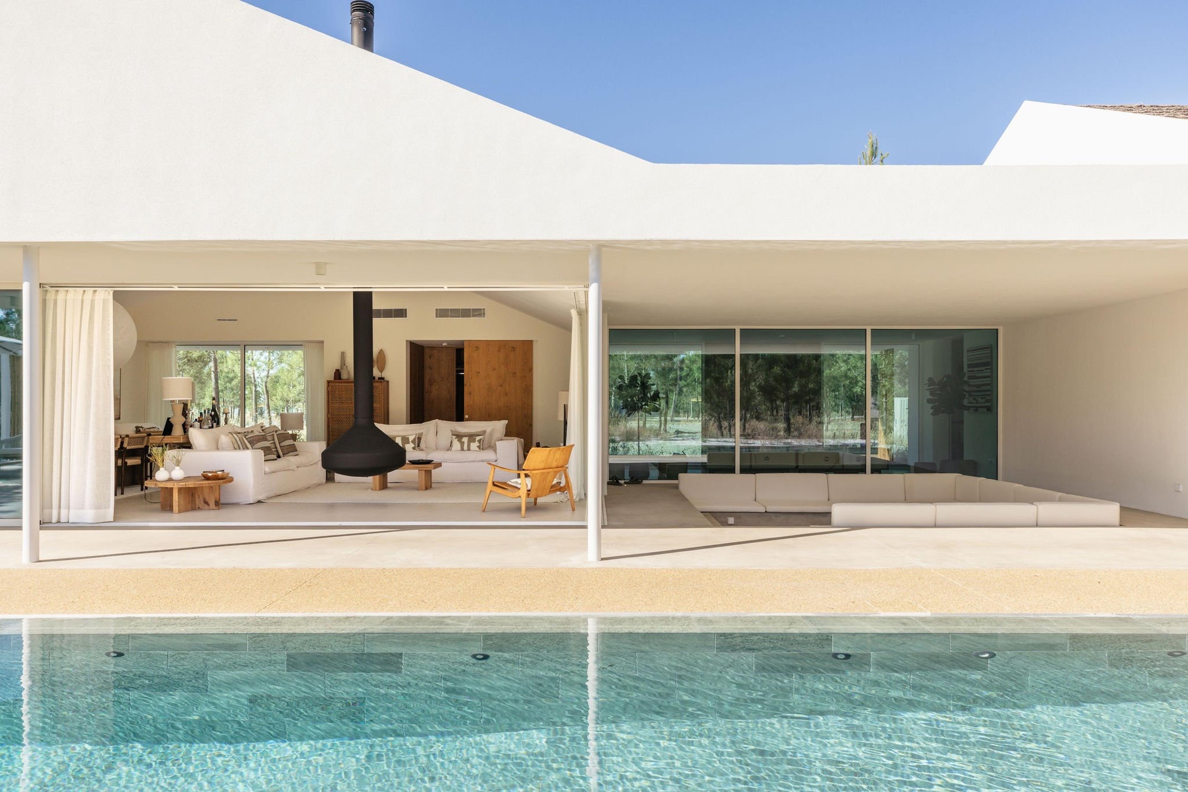 A swimming pool beneath an overhanging roof, with an open living room on one side and a built-in outdoor sofa on the other, trees reflected in the glass.