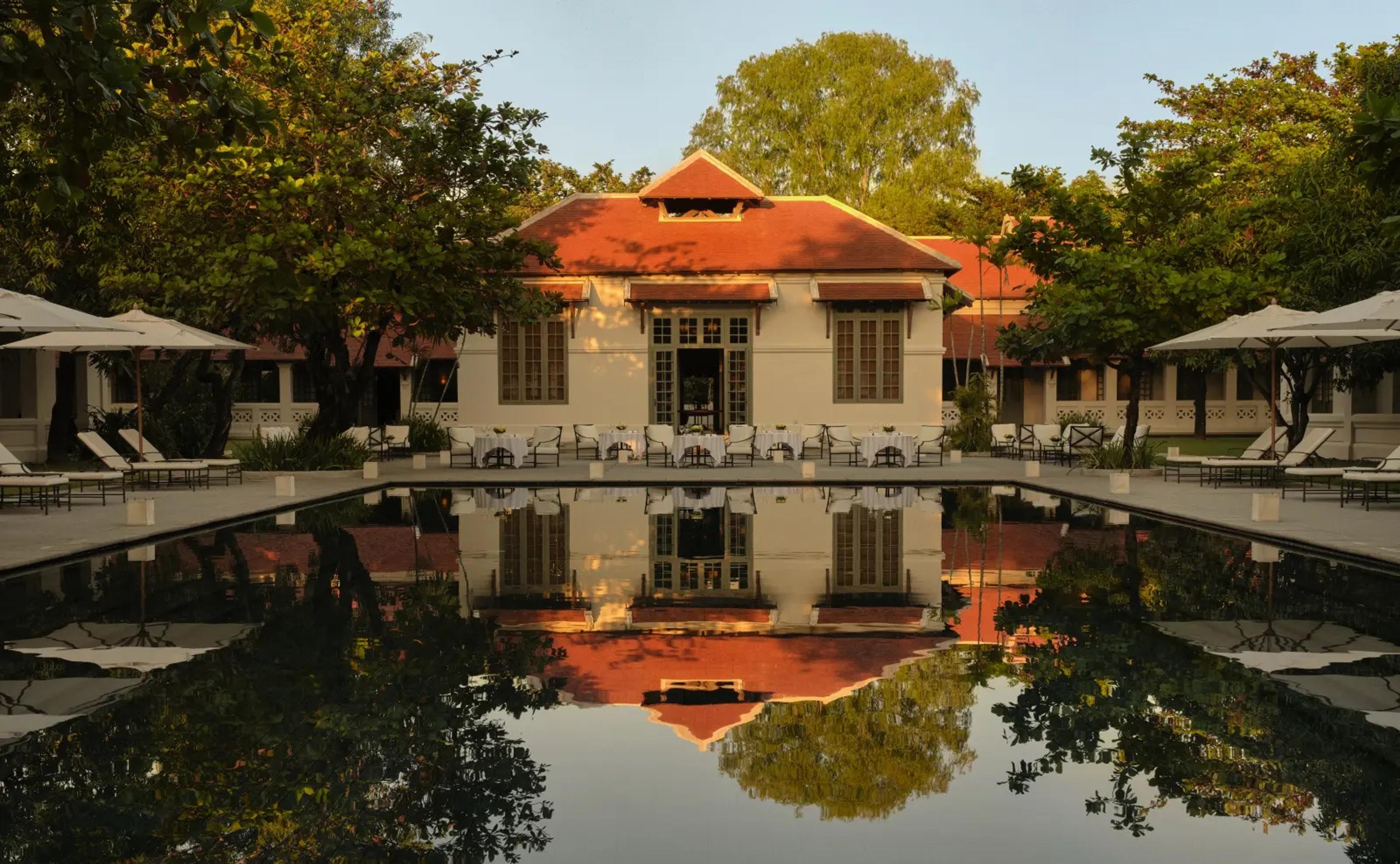 A long reflecting pool in front of a white colonial building with a steep red tiled roof, sun loungers and umbrellas on both sides, lit by late afternoon sun.