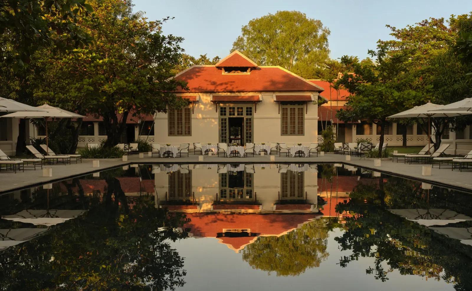 A long reflecting pool in front of a white colonial building with a steep red tiled roof, sun loungers and umbrellas on both sides, lit by late afternoon sun.