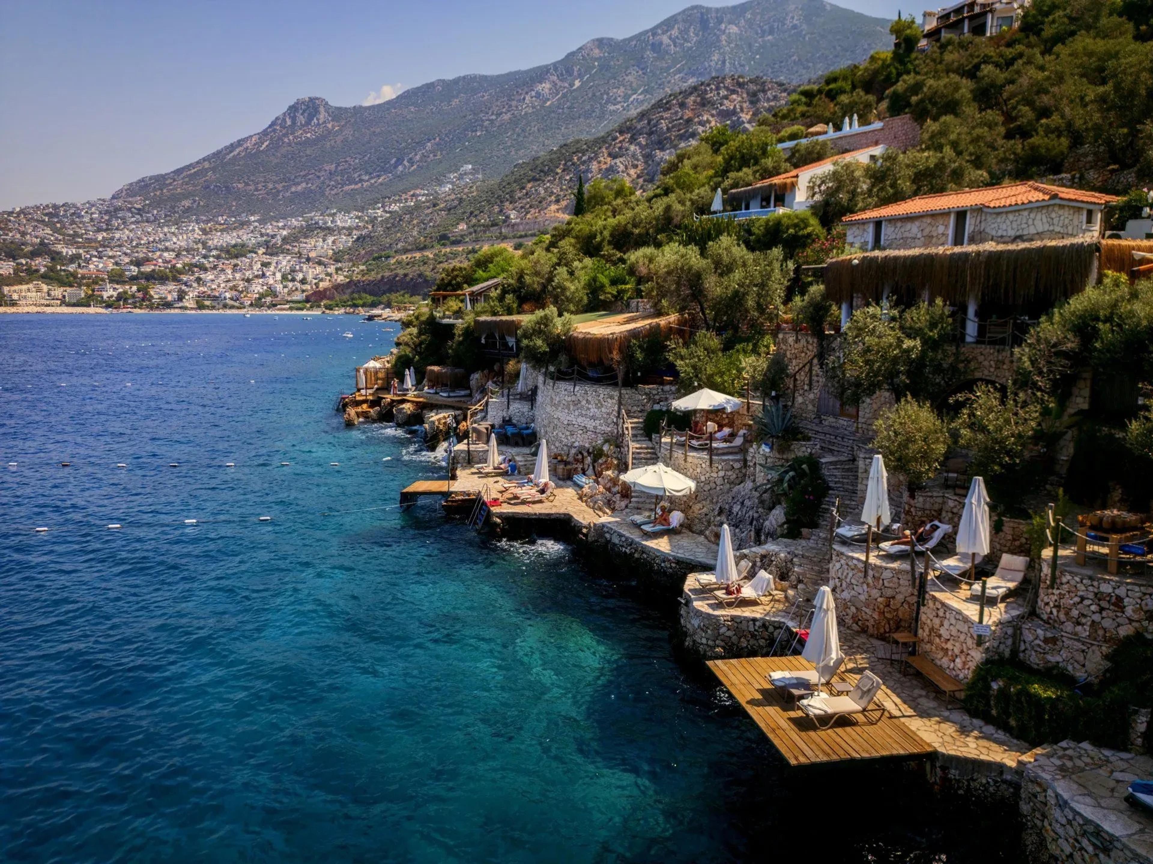 Aerial view of a beach club built into the rocks of a bay, with terraces, white parasols and cabanas, and a hillside town and mountain across the water.