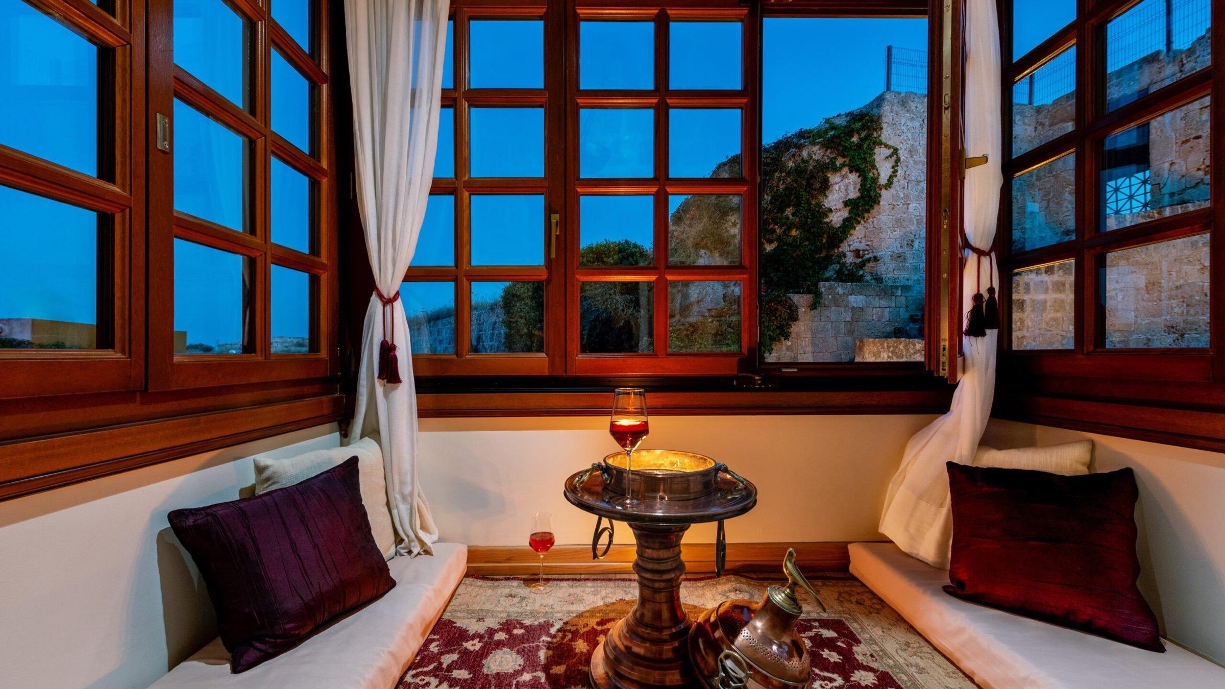 A window seat with cushions and a low table set with wine, overlooking an old stone wall at dusk.