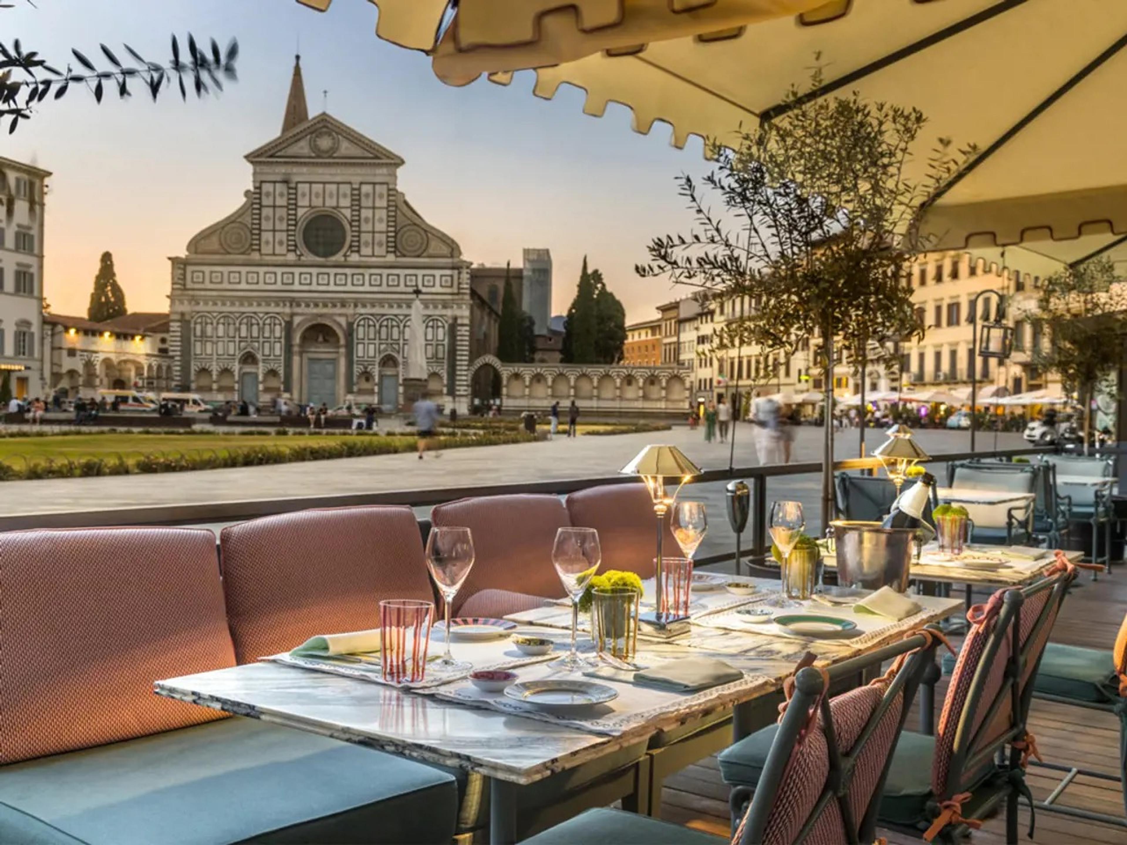 A rooftop terrace set for dinner with pink banquette seating and a laid table, looking across a lawn to the facade of Santa Maria Novella at dusk, with a few people walking in the piazza beyond.