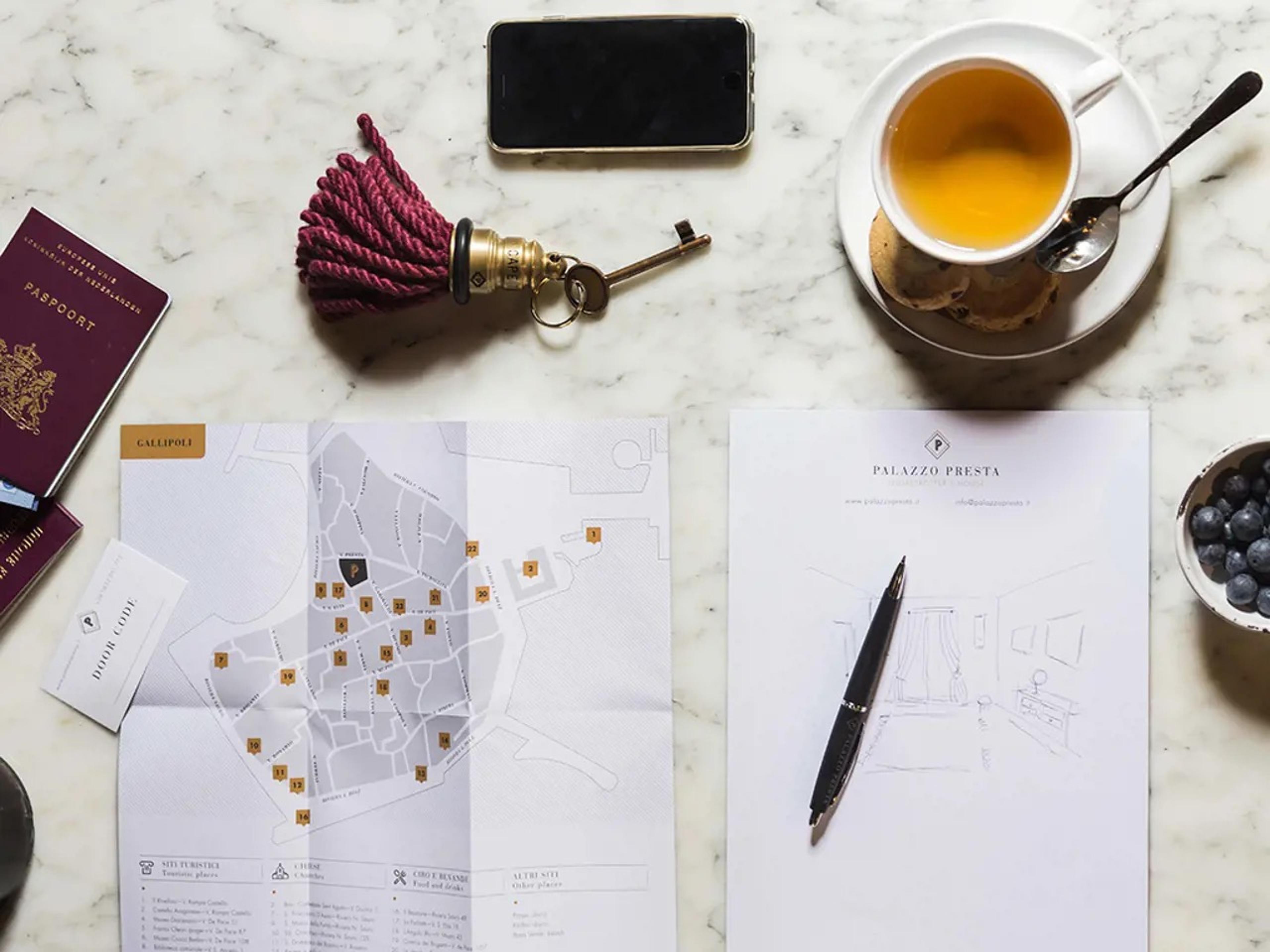 A marble table laid out with a passport, an old brass key on a tasselled fob, a cup of tea and biscuits, a folded map of Gallipoli, a notepad from Palazzo Presta with a pencil sketch of a room, and a bowl of blueberries.