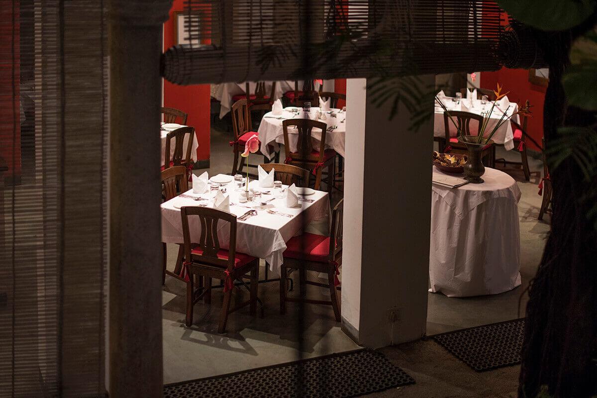 An empty restaurant courtyard set for dinner with white tablecloths and red-trimmed chairs, seen past a pillar and hanging blinds at night.