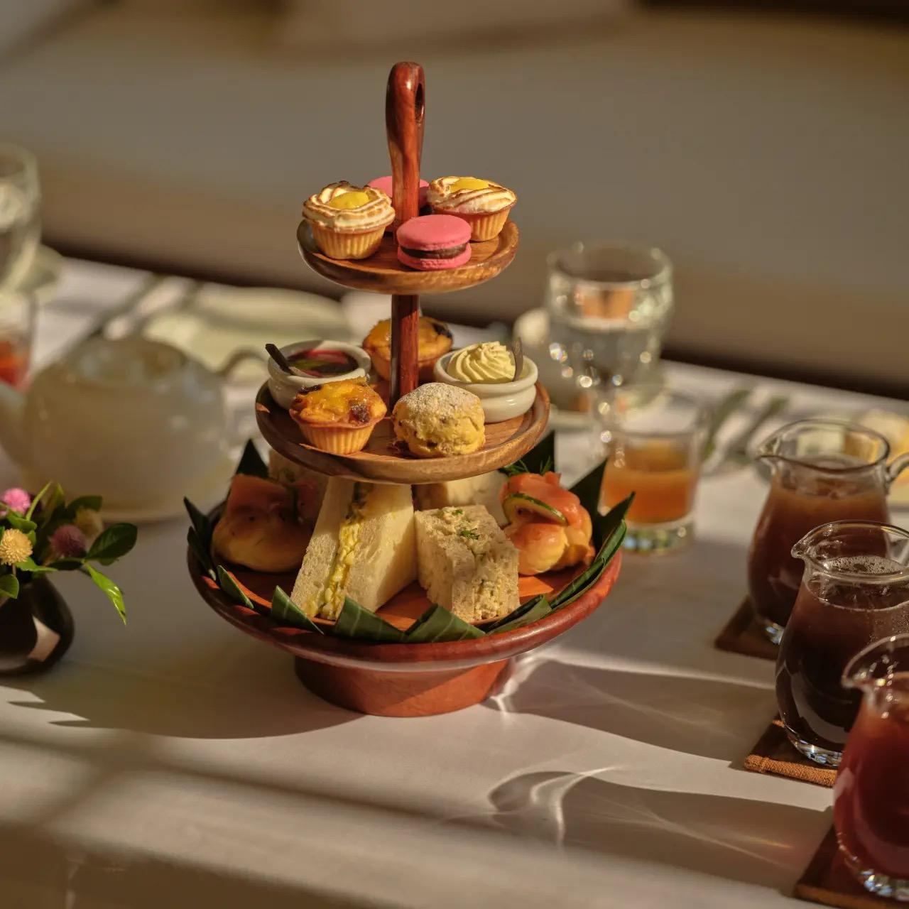 A three-tiered stand of pastries, scones and finger sandwiches on a table with jugs of iced tea in the sun.