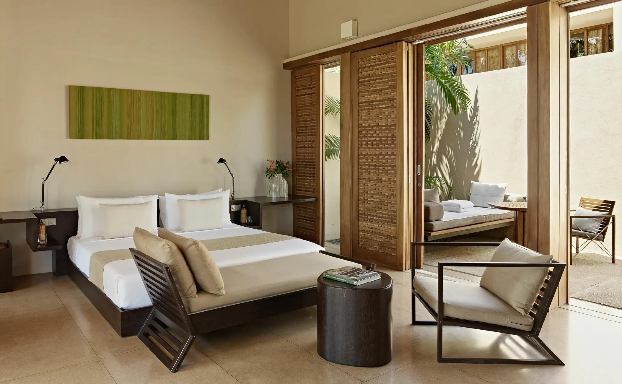A bedroom with a dark wood bed, cream pillows and a green abstract artwork, sliding wood slat doors open to a private courtyard and a daybed.