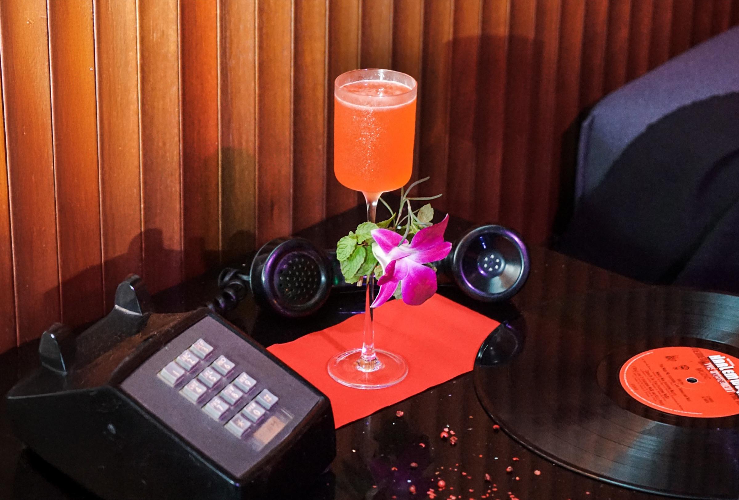 A cocktail garnished with mint and an orchid on a red cloth, beside a vintage telephone and a vinyl record on a dark wood table.