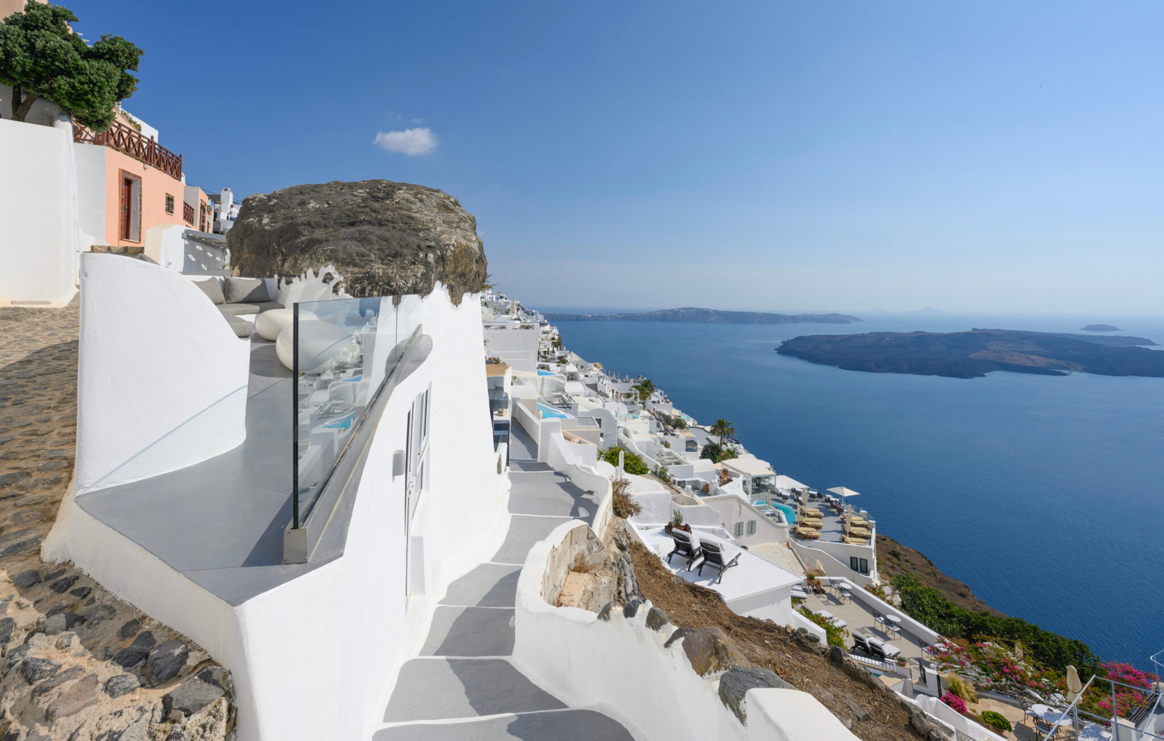 A row of whitewashed terraces and pools cut into a cliff above the sea, with the Santorini caldera stretching into the distance.