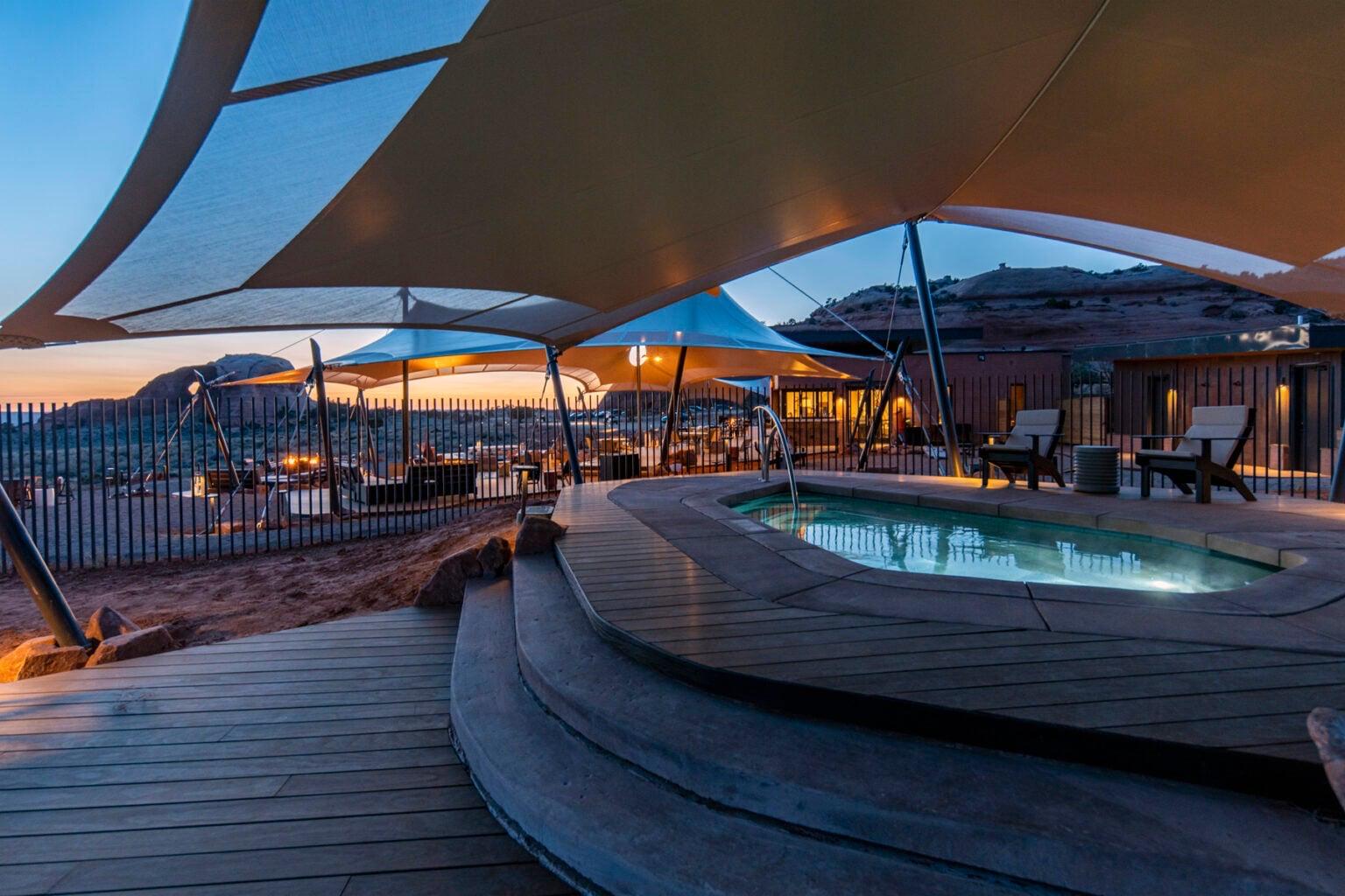 A curved timber deck around a small pool at dusk, with a large fabric shade sail overhead, lounge chairs beside the water, and the desert and rock ridges fading behind the railing.