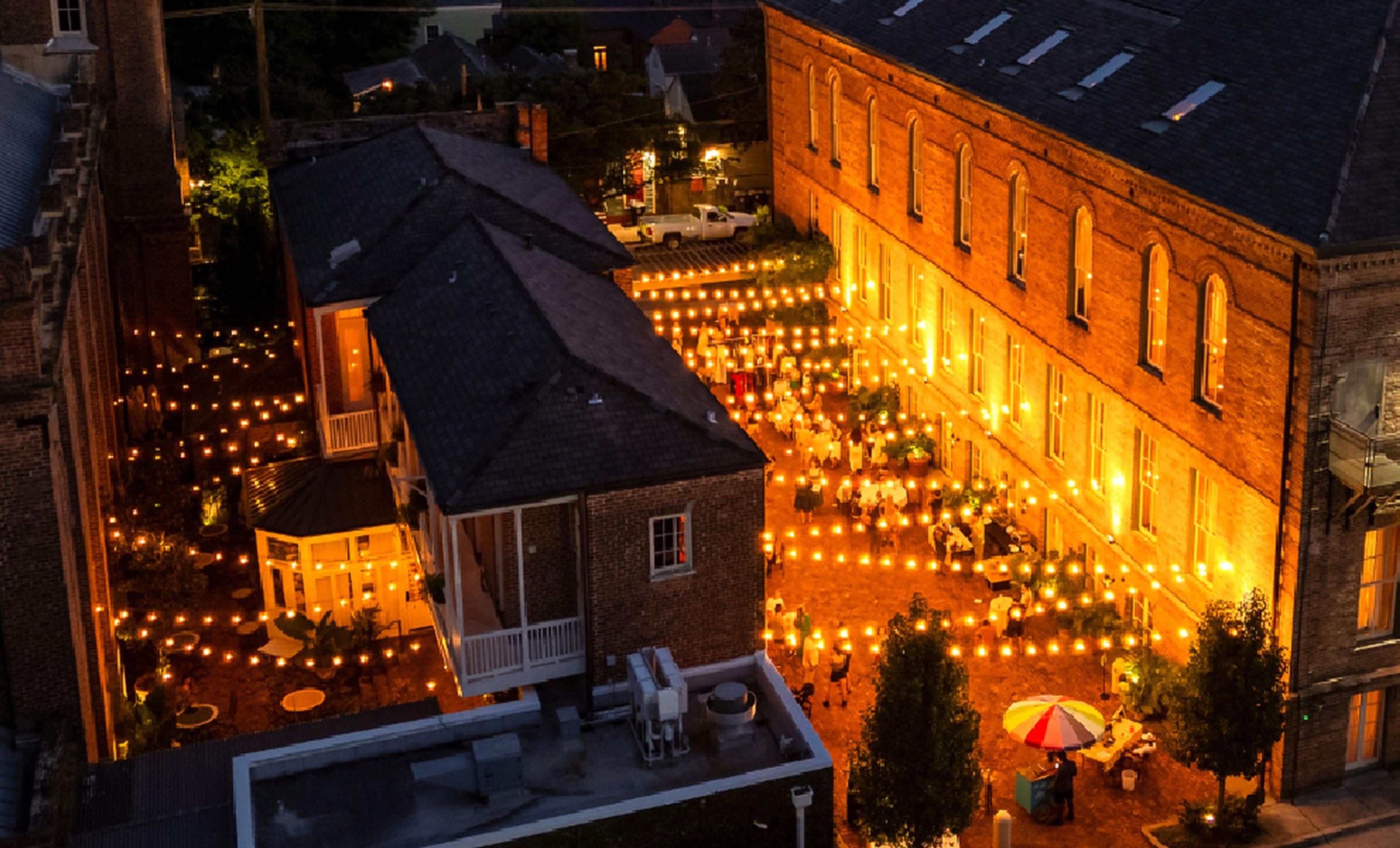 An aerial night view of a courtyard strung with lights between two brick buildings, long tables of diners below, and a striped umbrella in the corner.