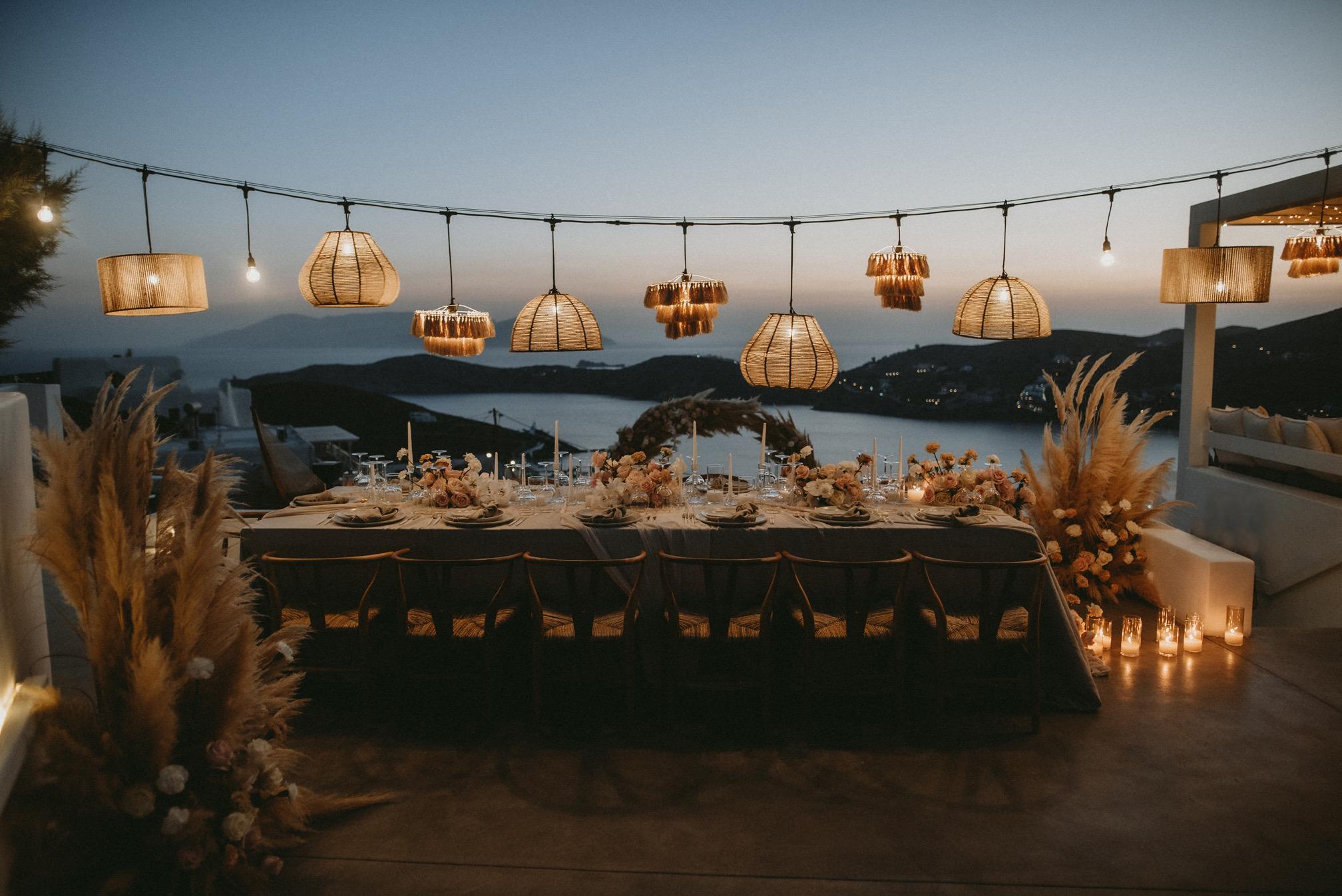 Tables on a terrace under strings of lights at dusk, the bay below